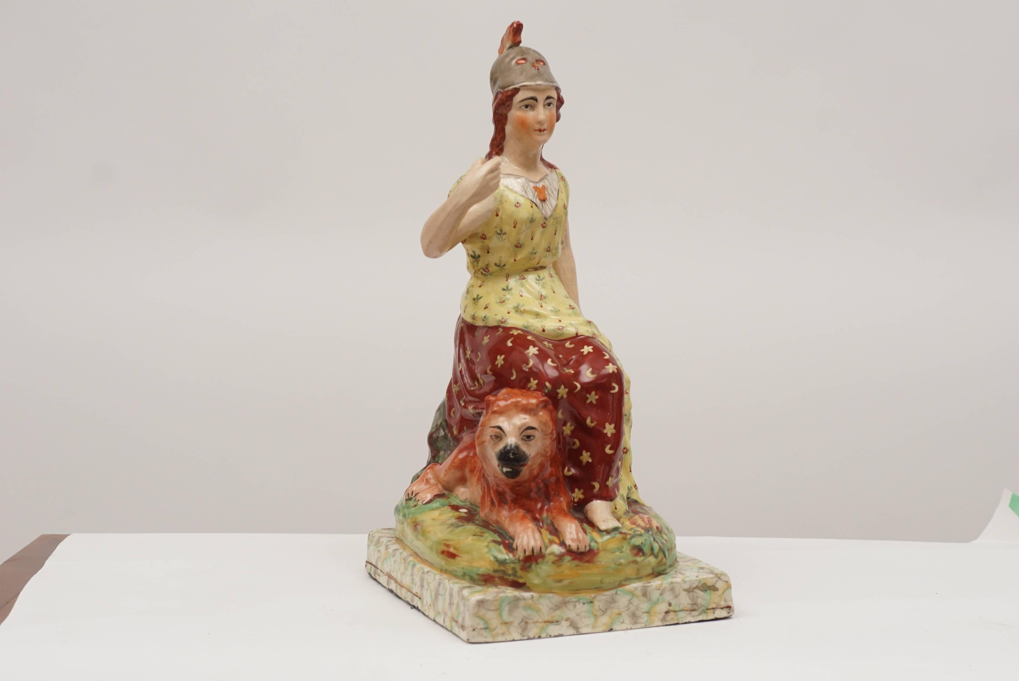 Allegorical Staffordshire pottery figure of a seated Britannia wearing a yellow and red dress. Imposing figure seated with her lion, circa 1820. Her shield is by her left hand. There is a repair to the tail end of her helmet. Overall she is quite is