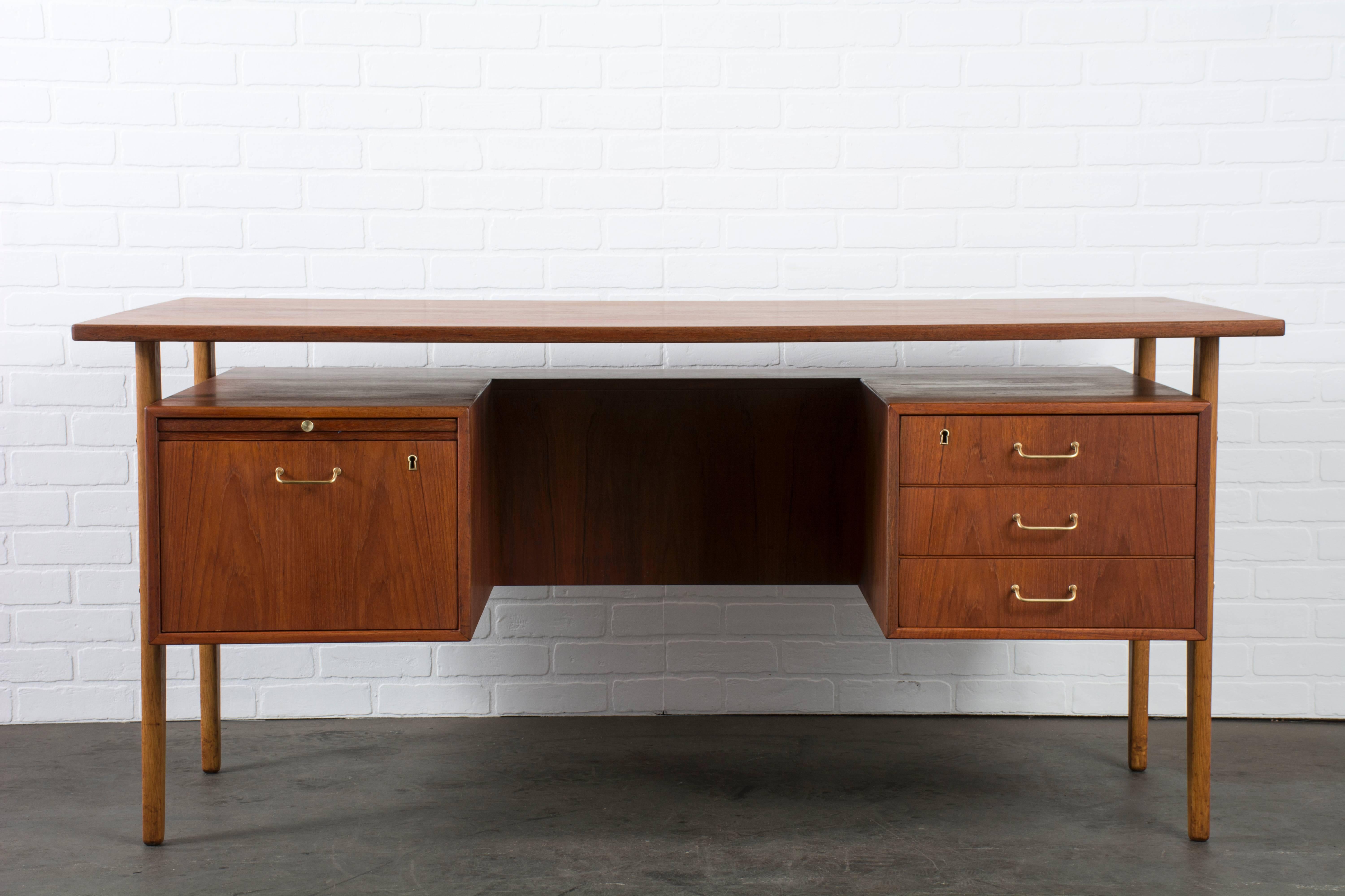 This vintage Mid-Century teak executive desk was designed by Torben Strandgaard in the 1960s. It features a floating top, a finished back with a shelf, three drawers on the right and one file drawer and pull-out tray on the left. Solid brass