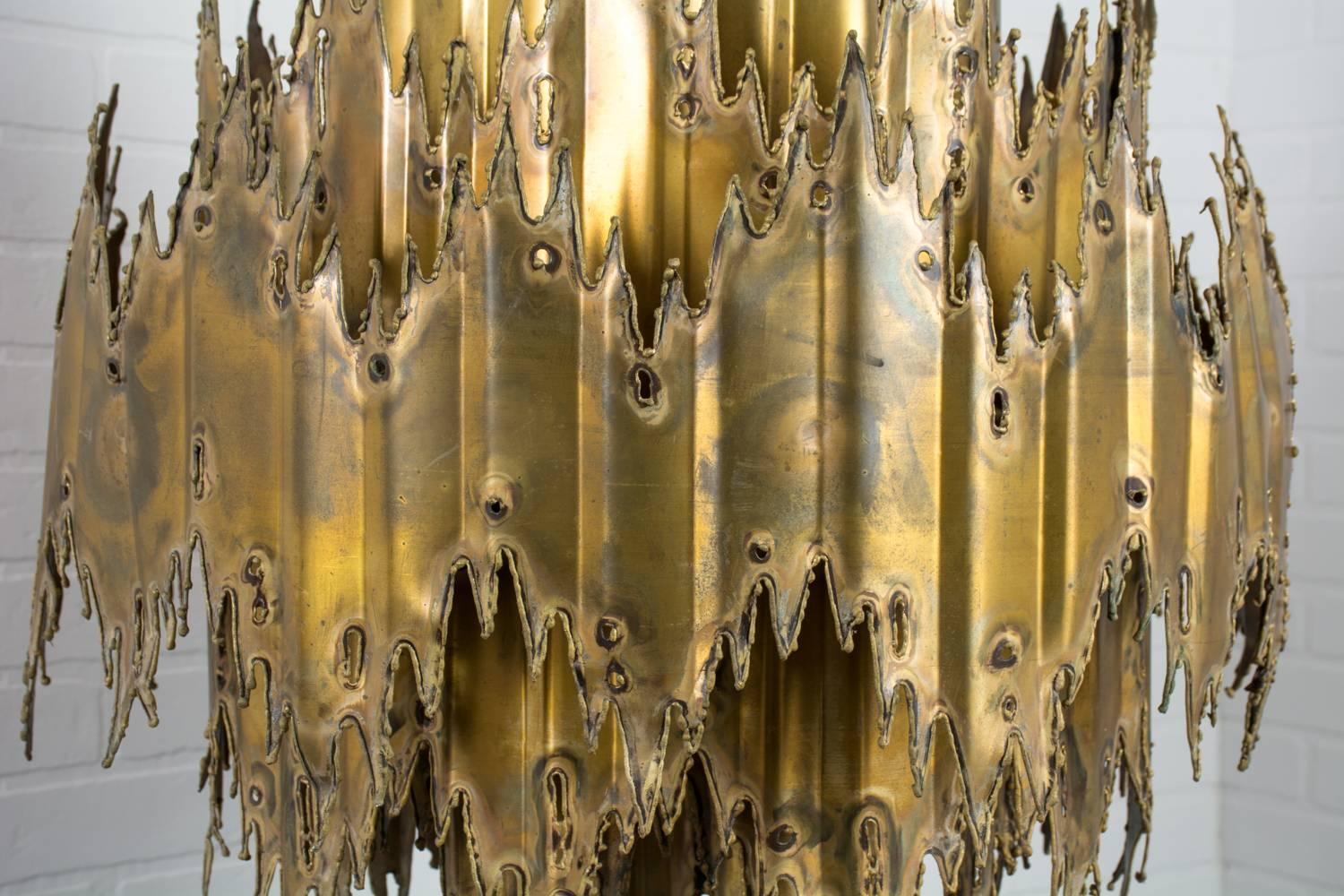 This striking Mid-Century brass chandelier was designed by Tom Greene for Feldman Lighting in the 1960s. It is constructed of four tiers of torch cut metal with a patinated brass finish. This hanging lamp holds six smaller light bulbs and one larger
