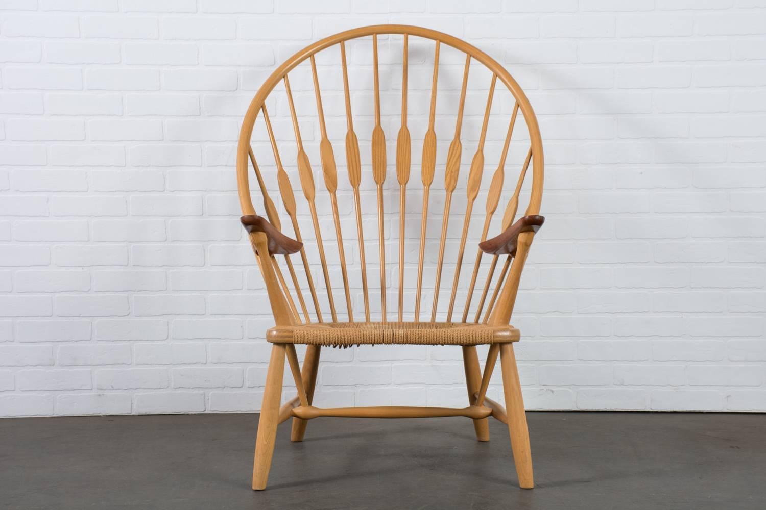 The 'Peacock' chair was designed by Hans Wegner in 1947 and manufactured by Johannes Hansen, Denmark. It features a solid ash frame with a rounded high back, contrasting teak arms, and a woven paper cord seat. 