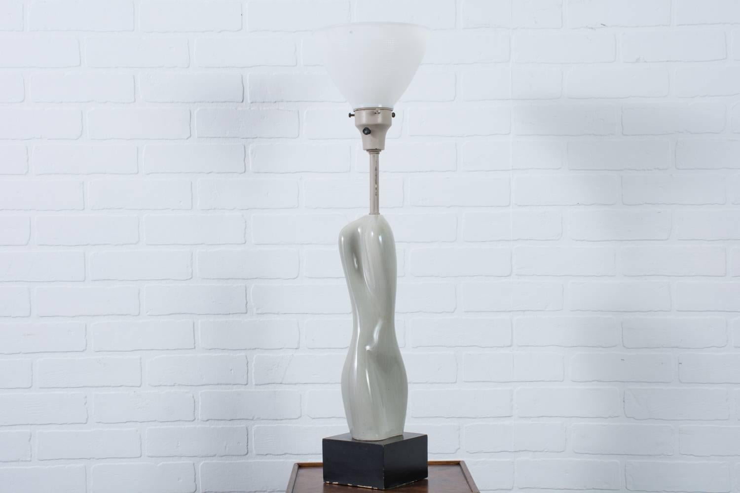 This vintage midcentury ceramic table lamp was designed by Russel Wright in the 1940s. The ceramic body of the lamp is in the form of a nude twisting to one side with a grey glazed finish. It is mounted on a black lacquered wood base and has a metal