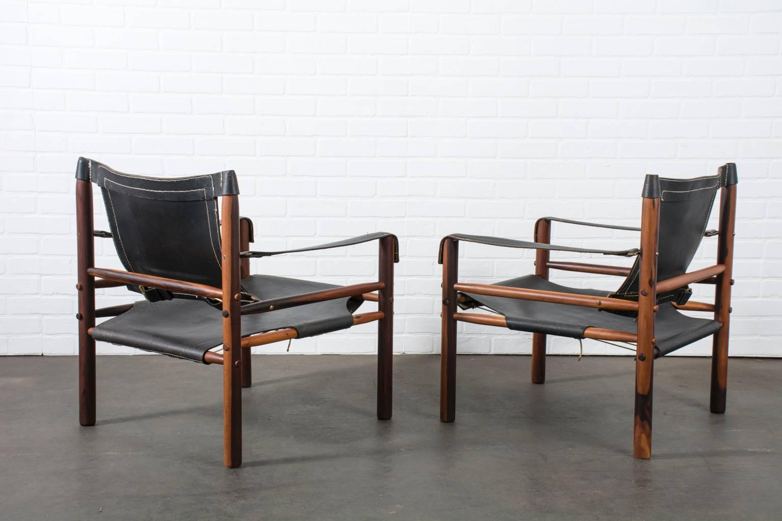 This is a pair of vintage Mid-Century safari chairs by Arne Norell. The have rosewood frames, black leather upholstery, and brass hardware.