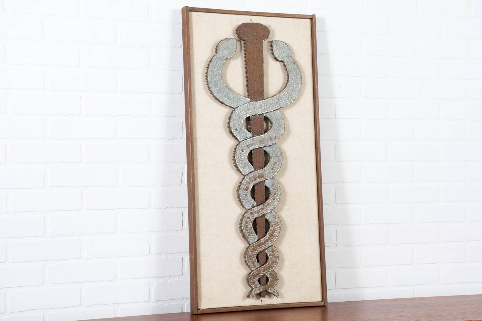 This vintage art piece consists of nails hammered into a linen covered wood background in the form of Caduceus, which is a rod entwined by two serpents (sometimes surmounted by wings) that was carried by the Greek god Hermes. In modern times it is