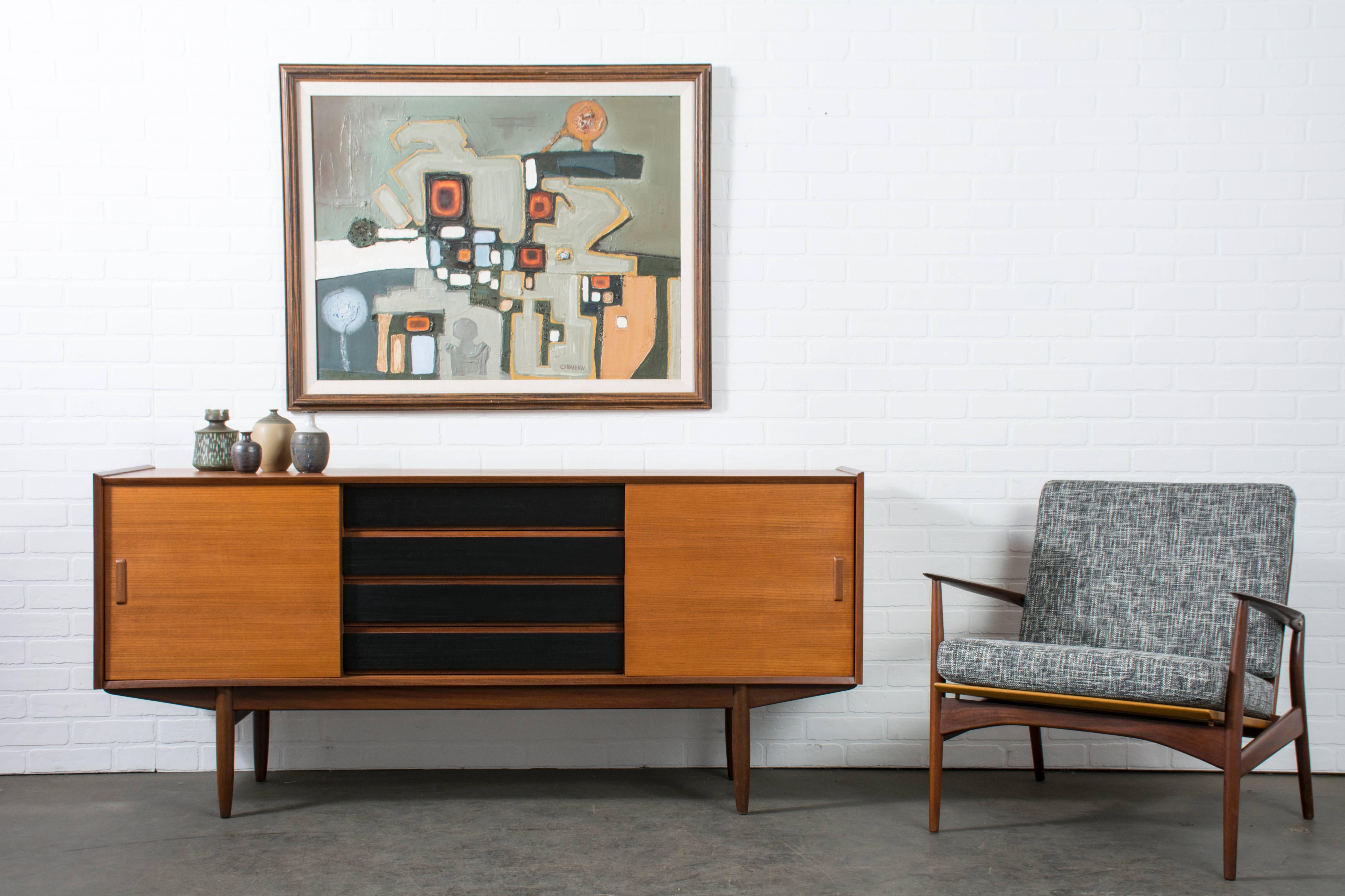 This Mid-Century Modern credenza is teak and walnut with black drawer fronts. It features four drawers in the middle (the top one with dividers) and one sliding door on each side that conceals one adjustable shelf. It is marked Hulmefa (Netherlands,