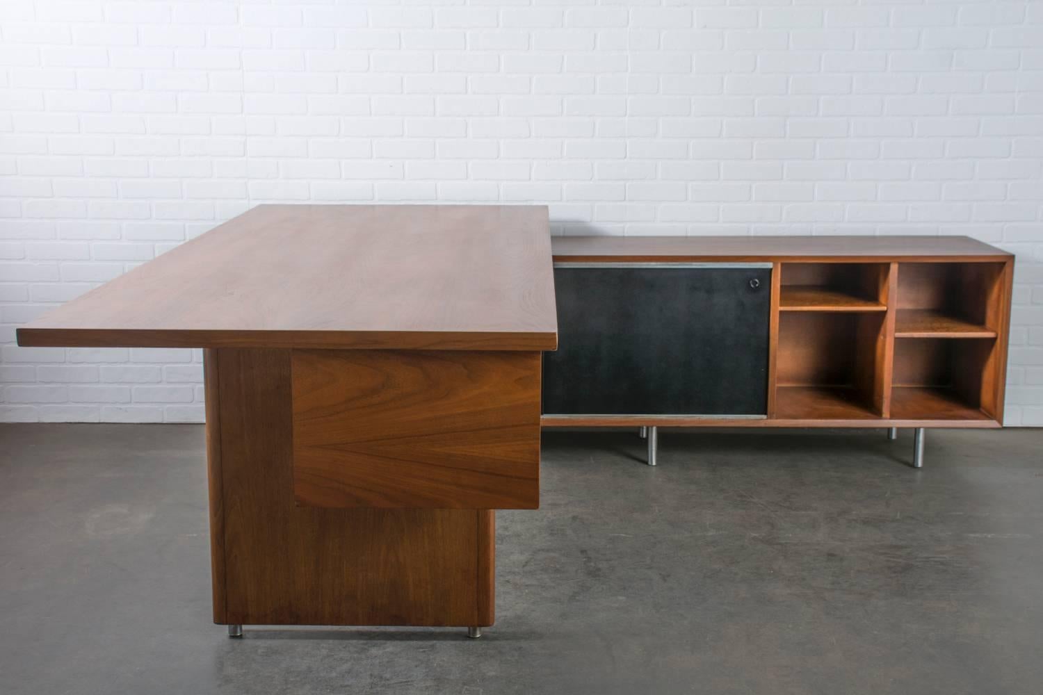 This Mid-Century Modern walnut executive desk was designed by George Nelson for Herman Miller in the 1950s. It features a large walnut desk top with three drawers and a credenza return with black sliding doors (on front and back), four drawers