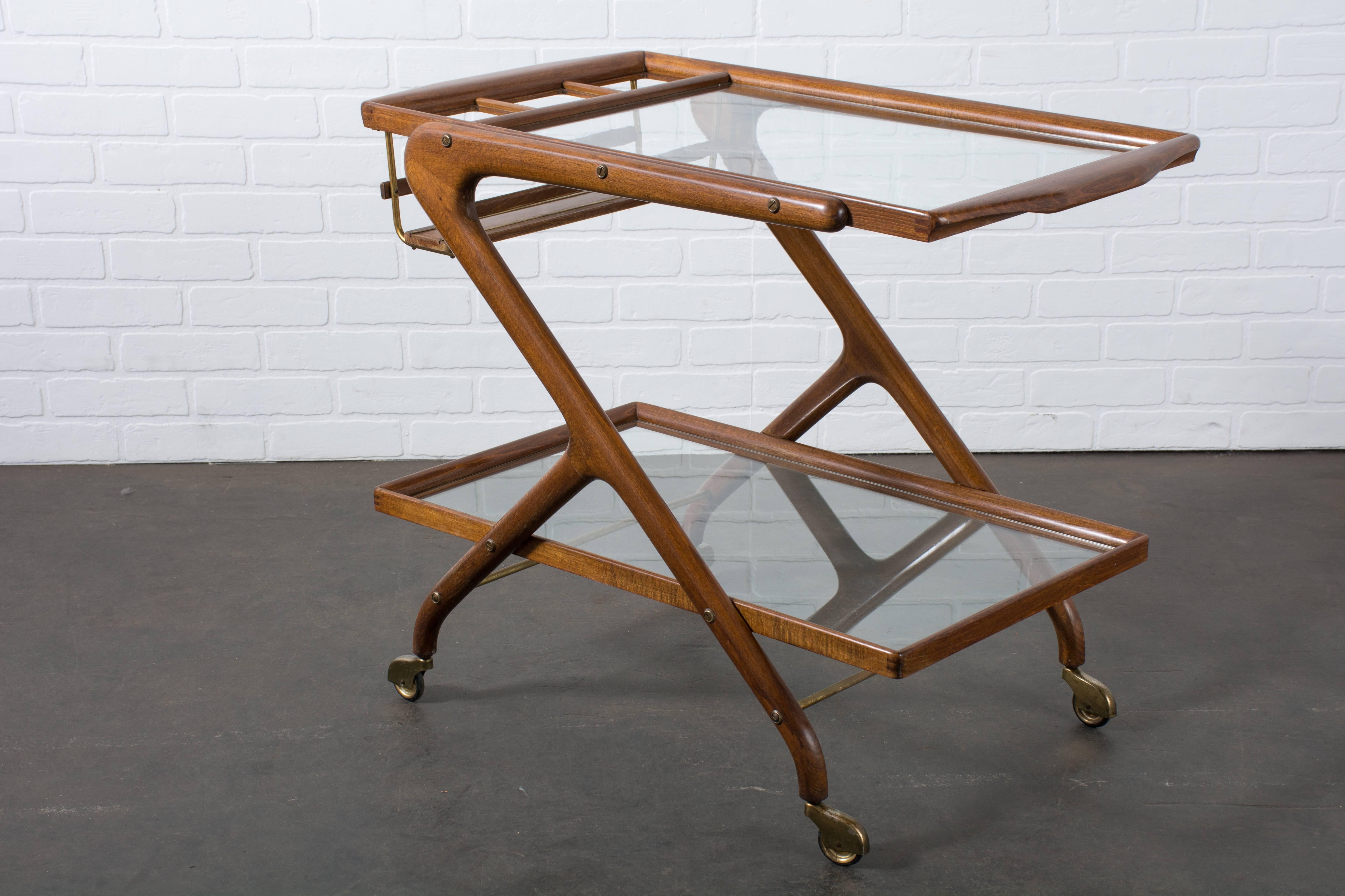 This vintage Mid-Century bar cart was designed by Cesare Lacca, Italy, circa 1950s. It features a sculptural walnut frame, two glass shelves, slots for bottles and metal details with a brass finish. This cart rests on four casters, so it can be