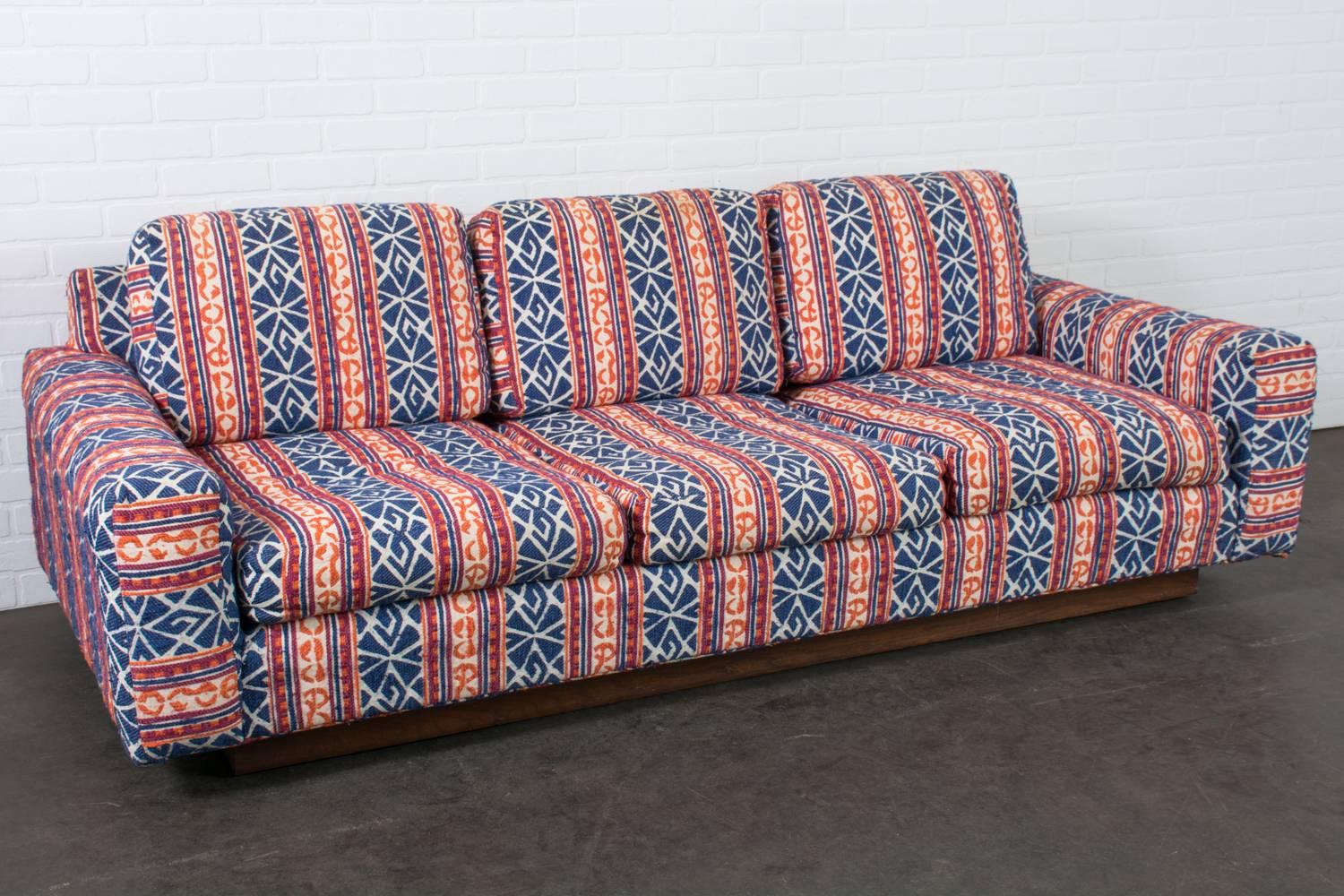 This low Mid-Century Modern sofa has a wood base with a walnut finish and is upholstered in a vintage fabric with shades of blue, orange, deep fuschia, and off-white, circa 1970s. This is the perfect vintage sofa for a modern Bohemian living room.