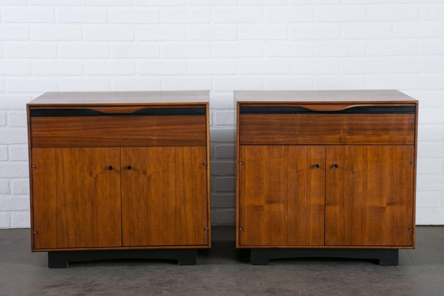 These Mid-Century Modern walnut nightstands were designed by John Kapel for Glenn of California in the 1960s. Each cabinet features one drawer and two doors that conceal a storage space with one adjustable shelf. The black details contrast