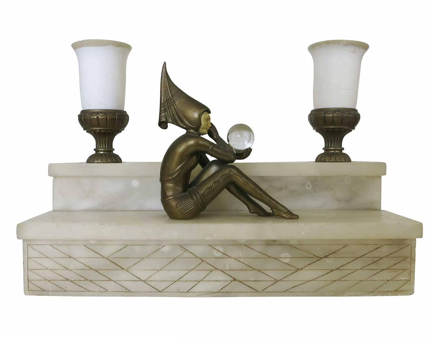 Large Art Deco alabaster mantel lamp featuring a bronze Harlequin figure with a pointed hood, looking into a crystal ball with two torchere style lights behind her.

 Also referred to as 