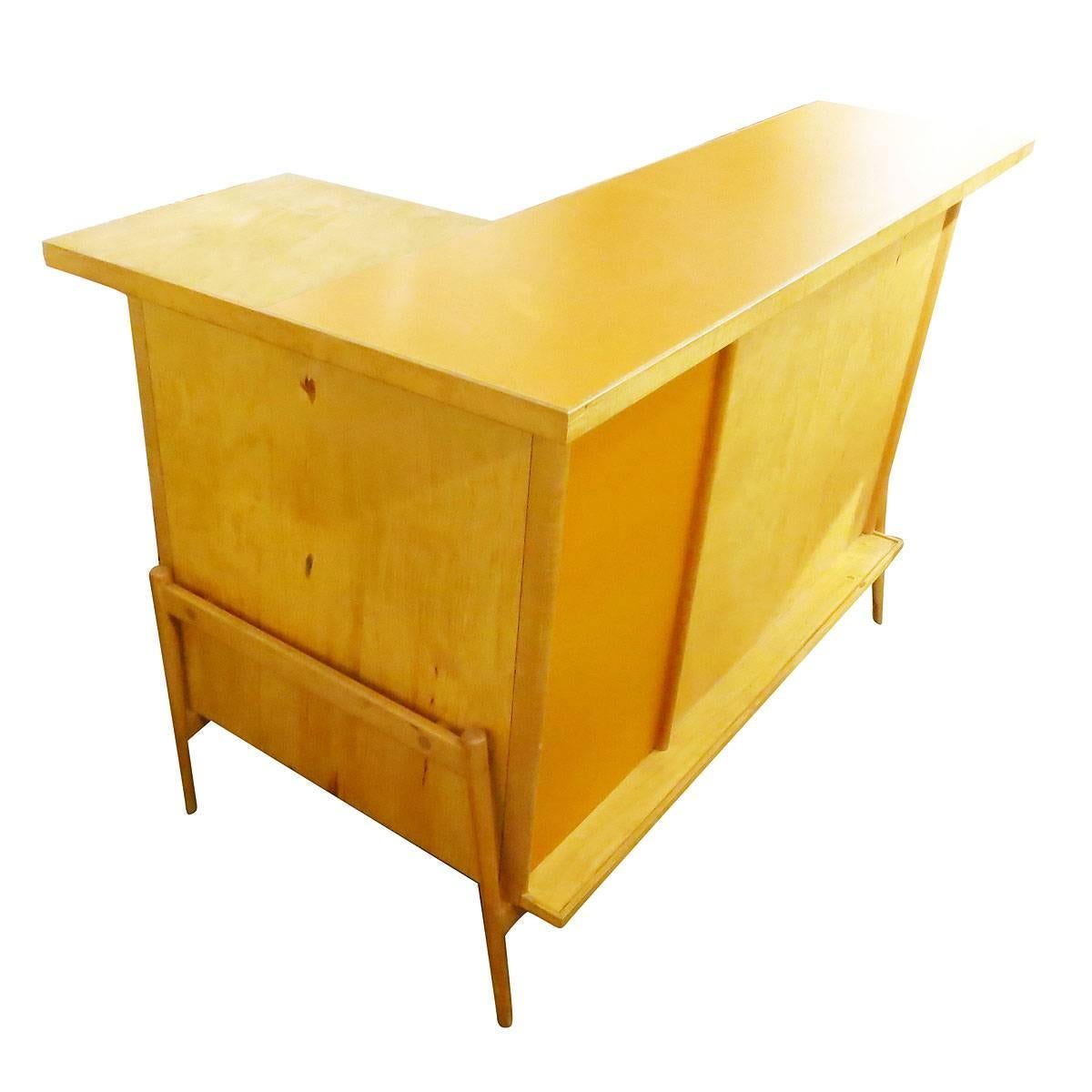 This Mid-Century Modern blond bent plywood bar circa 1950 comes with three matching bar stools. The Classic standing bar features an asymmetrical pale orange inset panel on the front. Behind the L-shaped unit there is plenty of storage for bottles