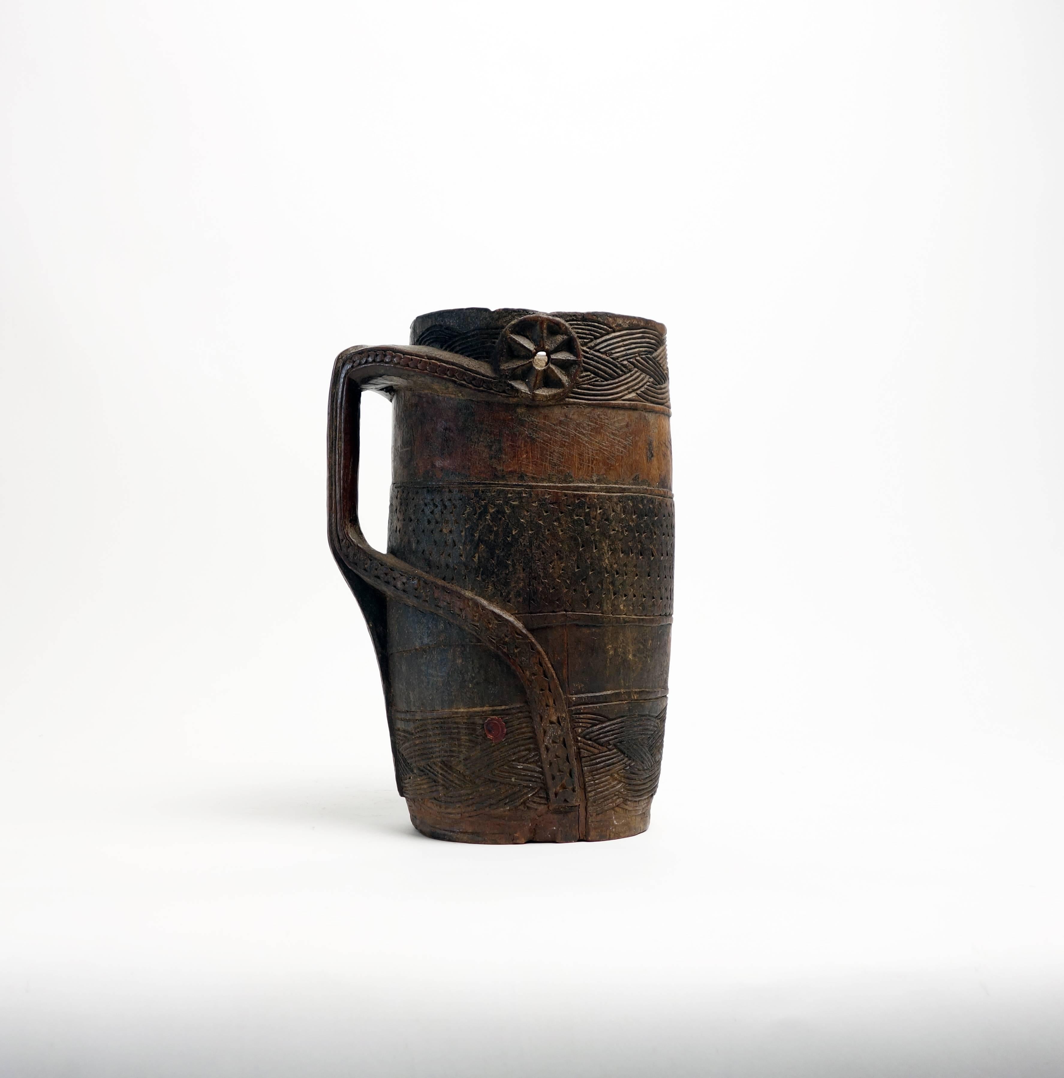 A ceremonial palm wine vessel of the Kuba peoples, carved in alternating horizontal bands of smooth wood and interlaced, abstract designs. The handle has an anthropomorphic quality. The Kuba take great care in the decoration of objects. Motifs are
