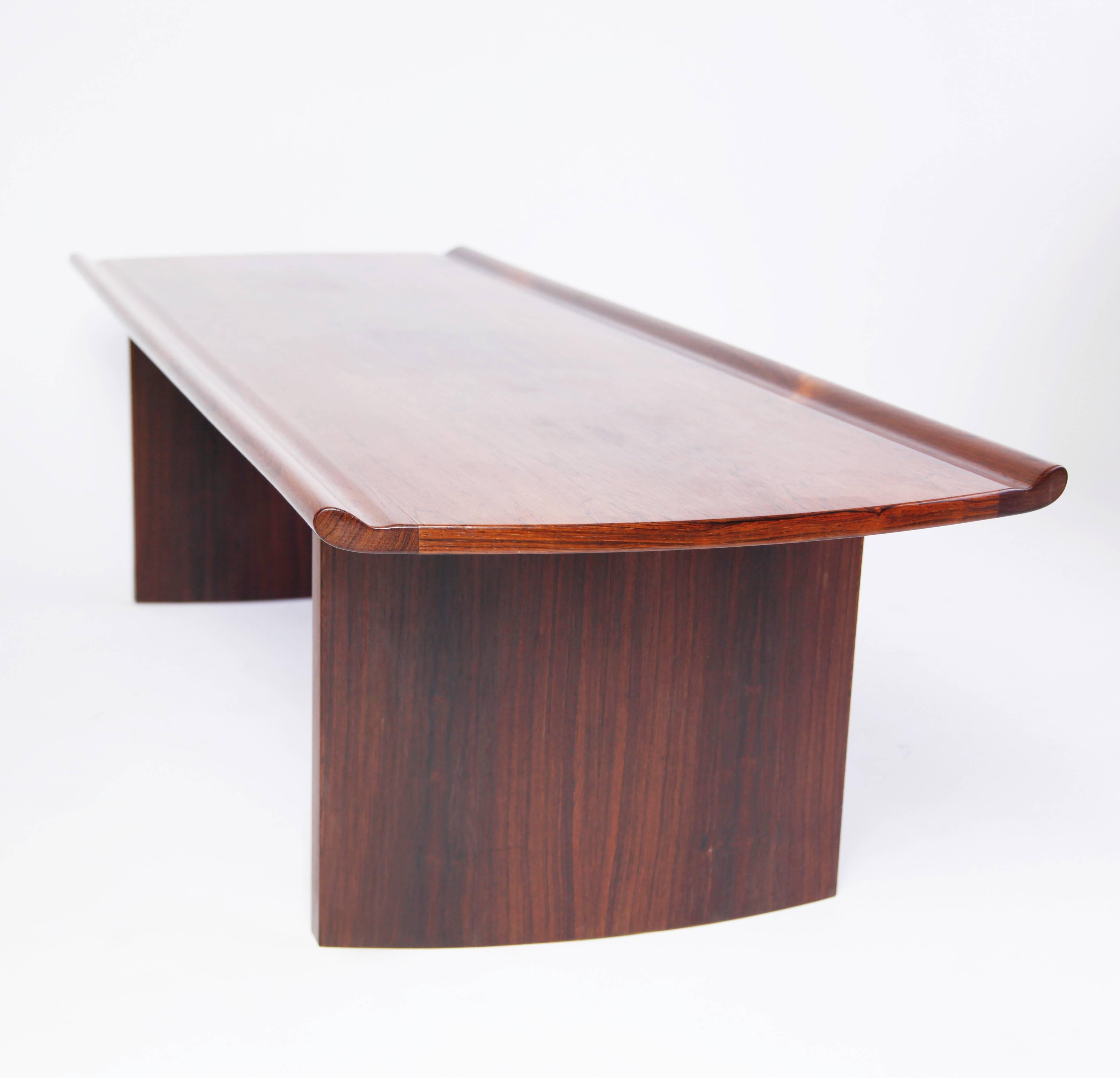 The top of this coffee table is solid rosewood, with an unexpected upward sweep. The legs are veneered rosewood, gently curving outward. A dramatic design, streamlined and modern, with an almost Japanese sensibility.