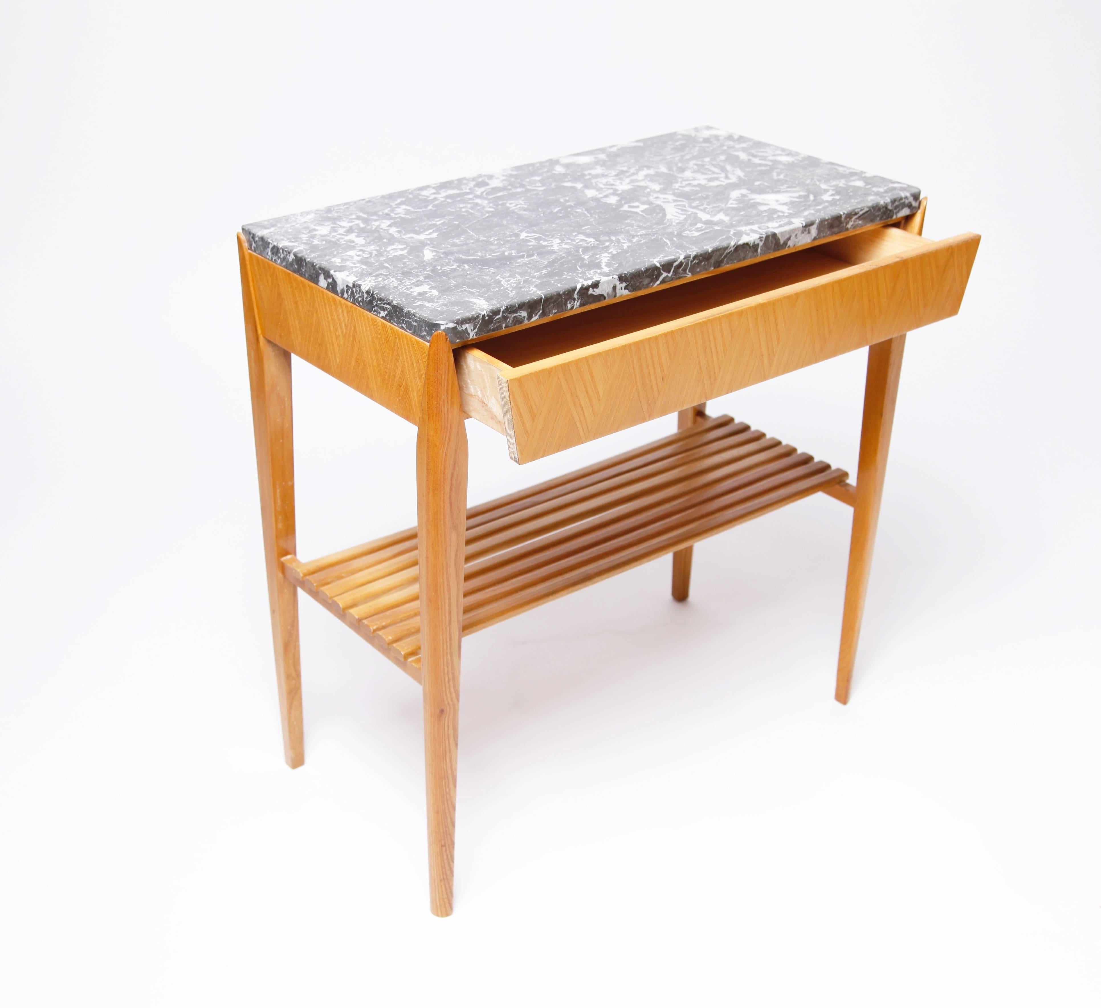 At first glance a very simple design, this small side table rewards closer examination. The dark gray and white marble top makes a nice contrast with the warm finish of the wood. The single drawer and the apron are ornamented with a very pretty