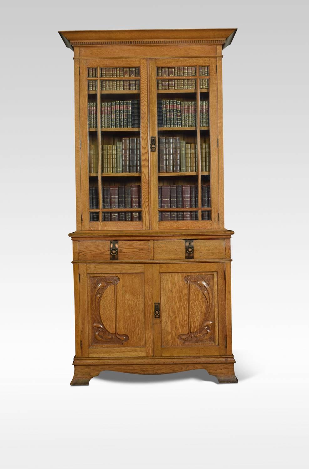 Art Nouveau oak bookcase, the projecting dental moulded cornice above a pair of glazed doors opening to reveal three adjustable shelves. The base section fitted with two short drawers and a pair of cupboards below with carved Art Nouveau panels and