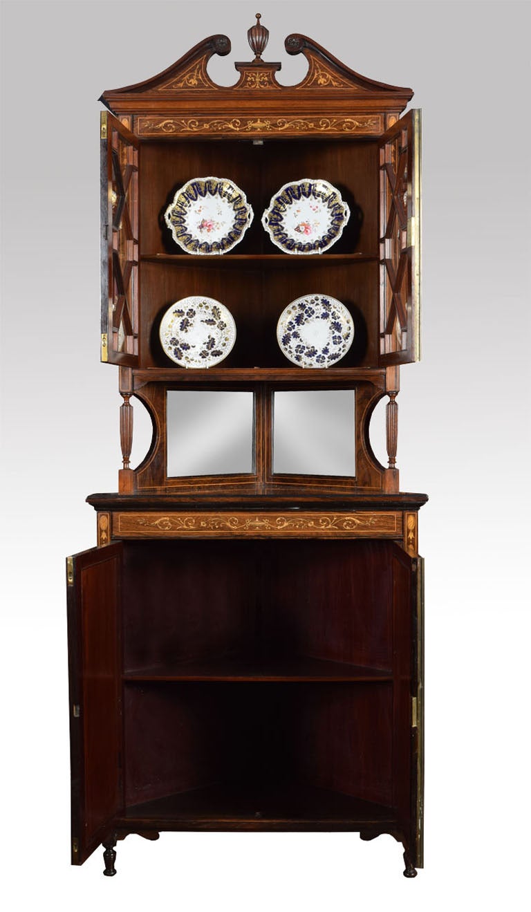 Edwardian rosewood inlaid corner display cabinet the arched swan neck pediment with central urn above two astragal glazed doors opening to reveal single fixed shelf above bevelled mirror centre. The base having two rosewood inlaid panelled doors