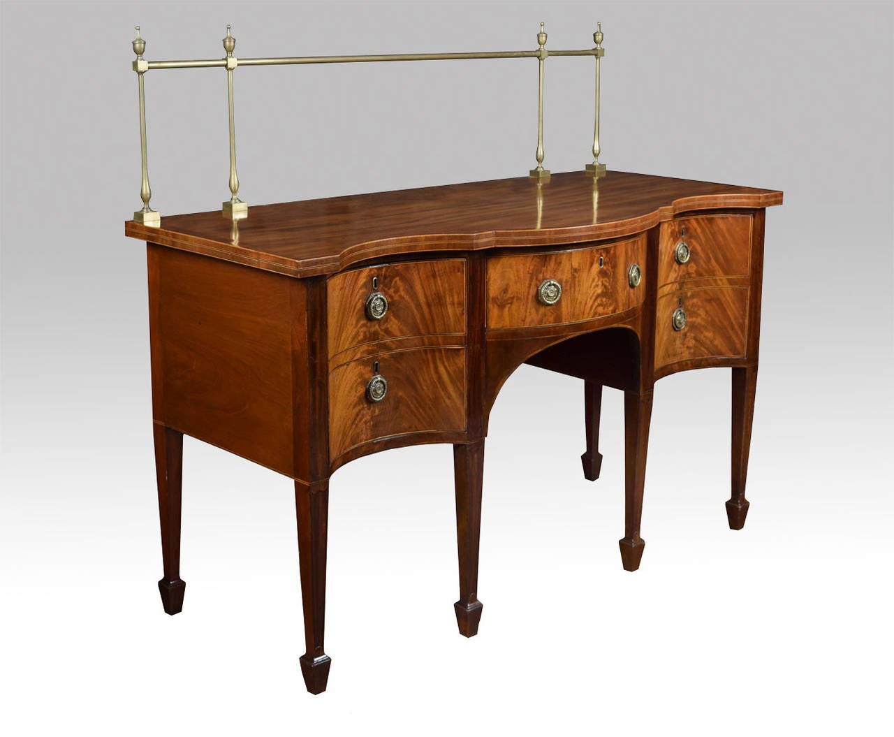 A mahogany sideboard having satinwood crossbanding, brass rail surmount having urn finials on baluster turned supports, the deep boxwood bordered top raised above single frieze drawer with brass ring handles, flanked by two deep drawers (one