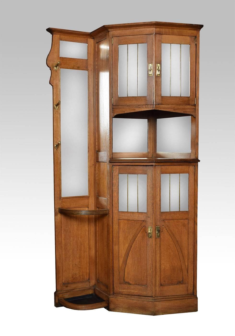 

Arts and Crafts oak corner hallstand the angled cupboards enclosed by mirrored panel doors separated by an open recess the opposing side with mirrored top having coat hooks and stick stand below all raised up on Plinth