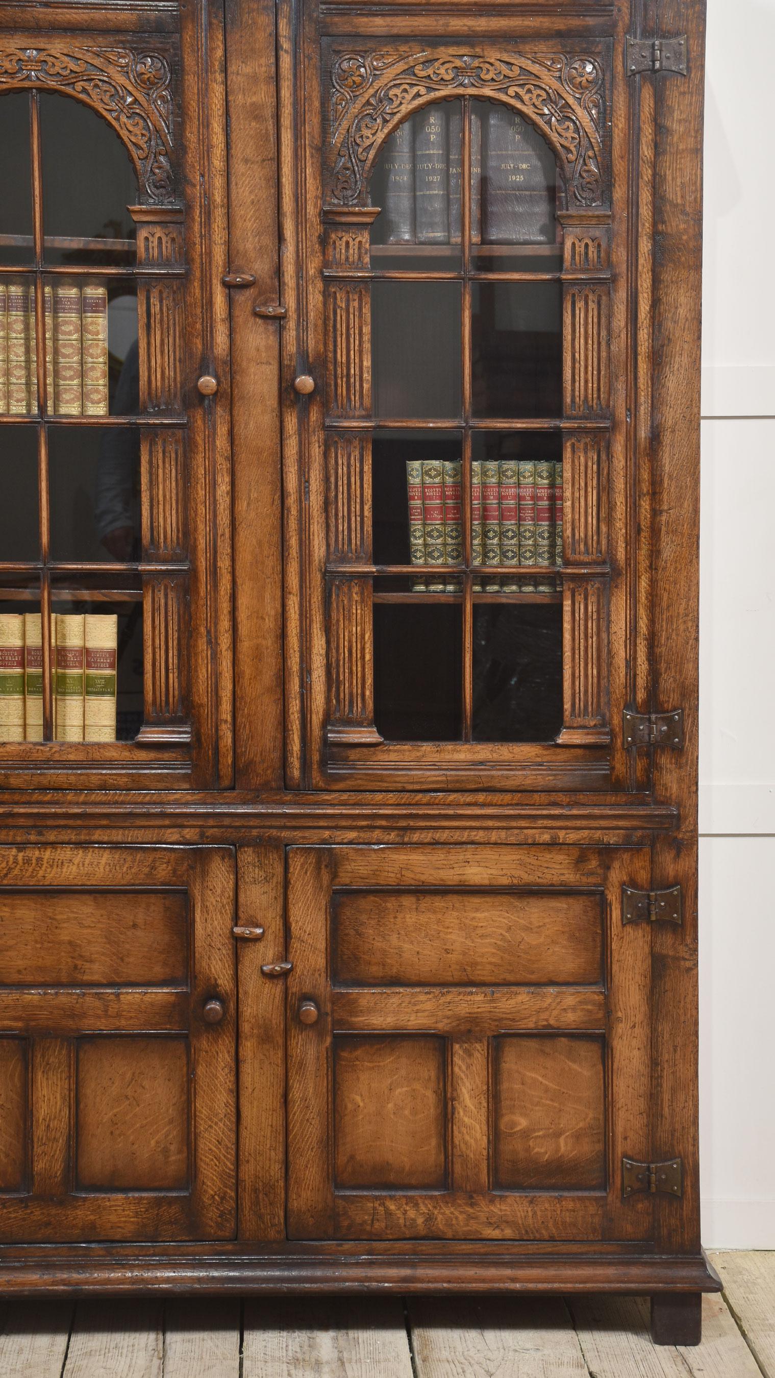 Carved oak bookcase having moulded cornice above two large arched glazed doors opening to reveal a shelved interior. The base section having a pair of panel doors opening to reveal further shelved interior. All raised on block