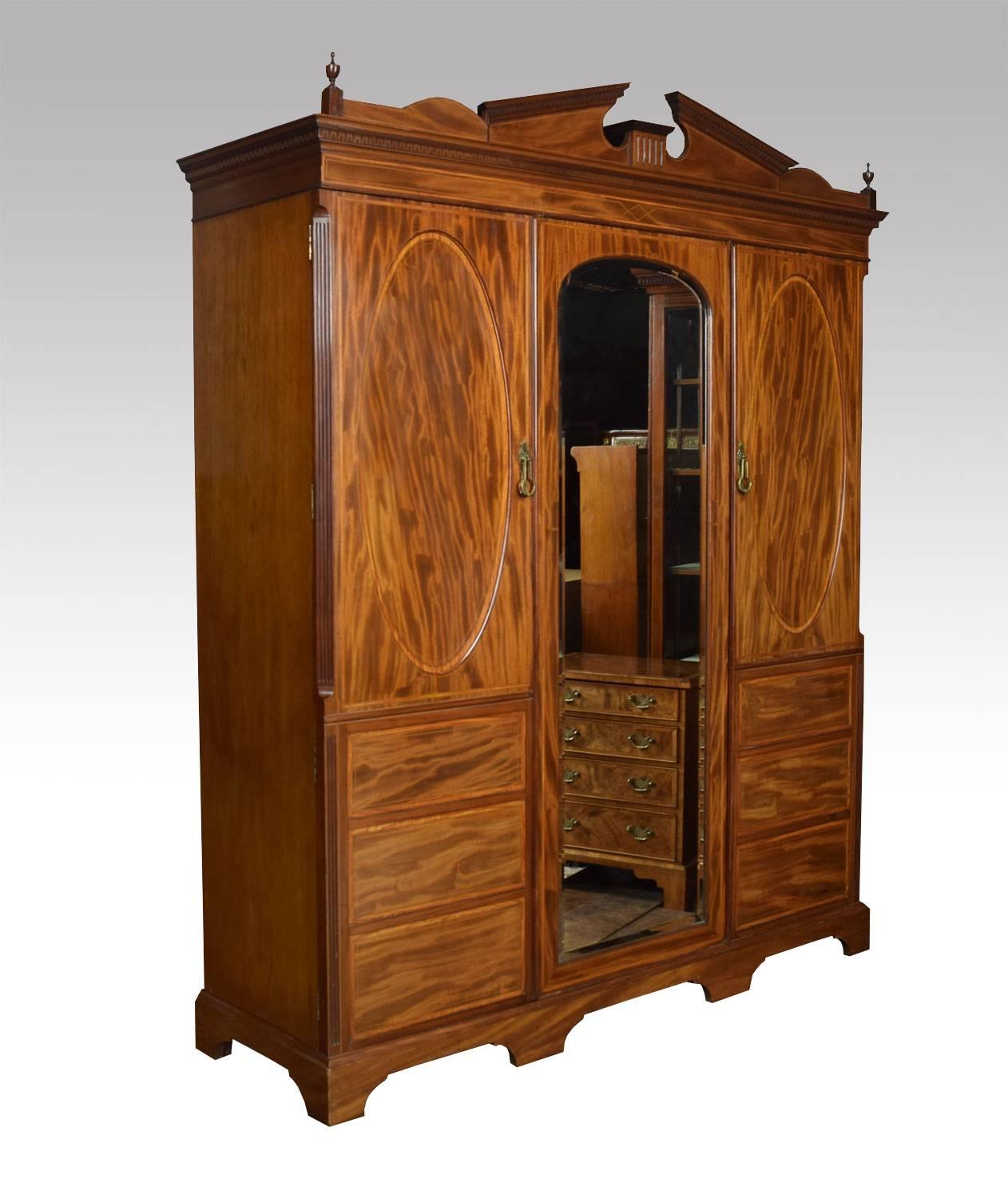 Figured mahogany three-door wardrobe the architectural cornice, flanked by urn finials above three large mahogany doors. The centre door with original full length mirror plate, surrounded with well figured oval panelled side doors having canted