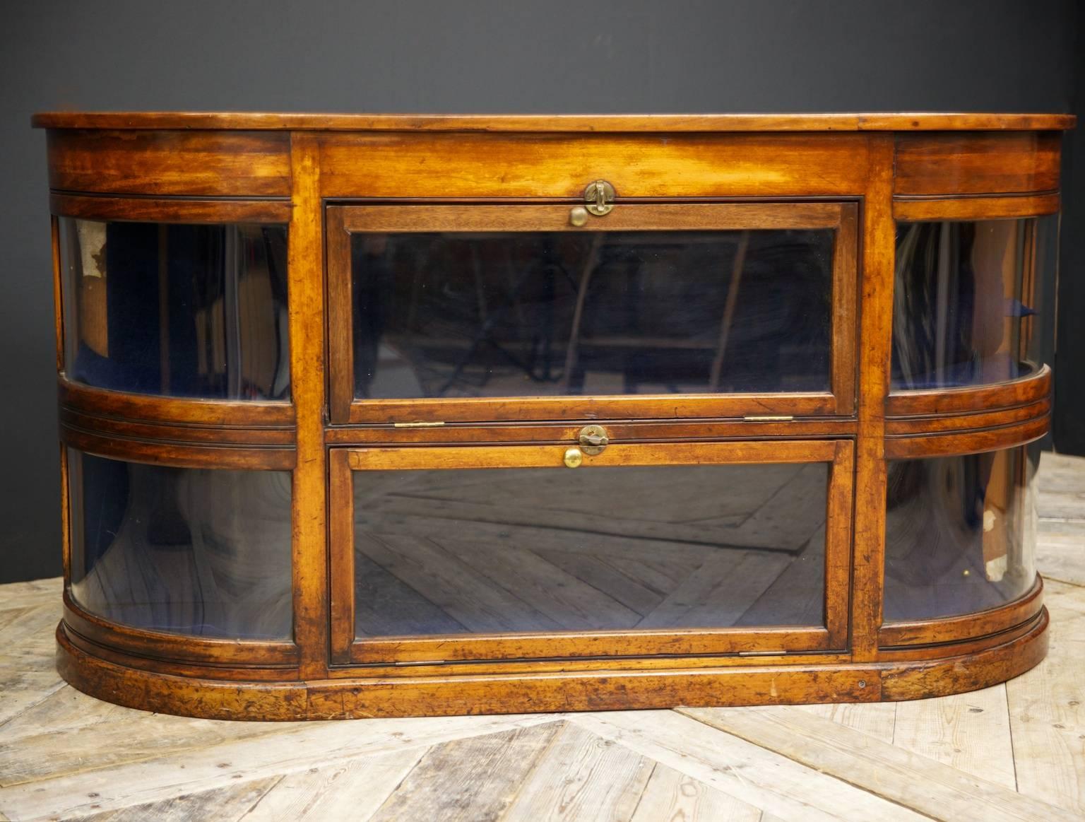 A glazed mahogany retail display credenza.

Curved glass on both sides, two drop-down fronts with locking bronze stays.

Very good original condition, the purple velvet liner pulls out to show bare pine lining.

English, 1920s. 