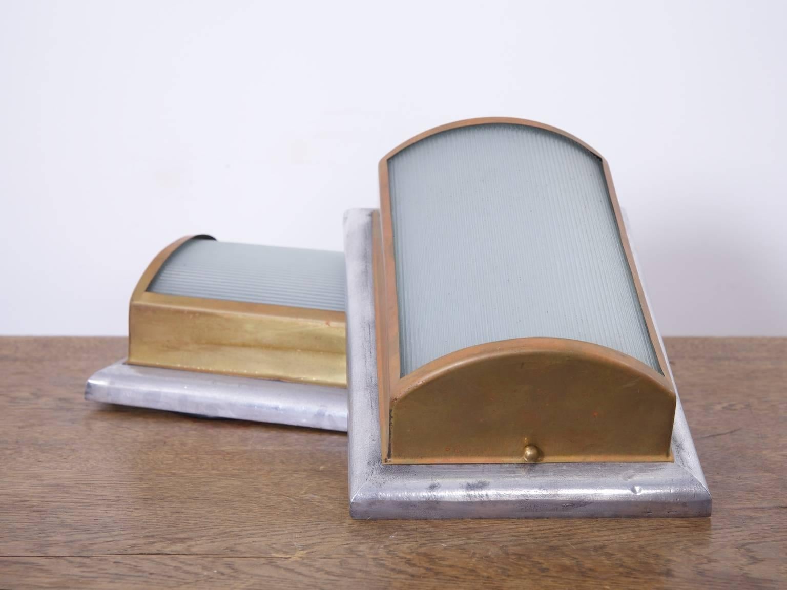 A pair of wall lights in the Art Deco taste.

The brass frame with curved Holophane prismatic glass lenses mounted on a cast alloy surround,

English, 1920s.

Rewired and pat tested.