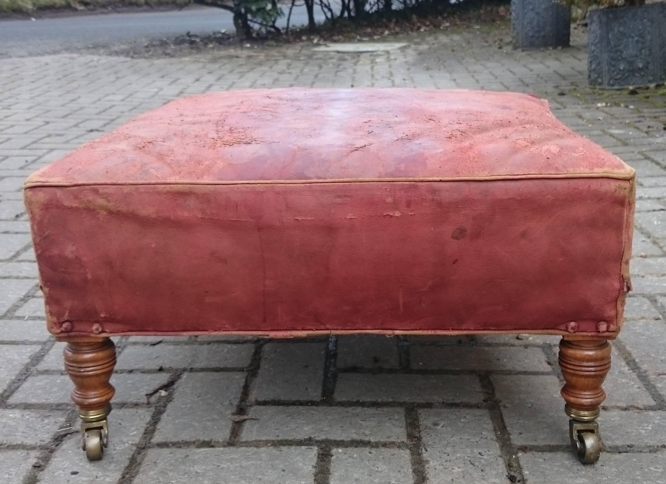 Upholstered footstool made by Howard and Sons of London standing on the turned legs typical of the company and upholstered in soft old red leather which might be the original. The casters are stamped and there is part of the original Howard label on