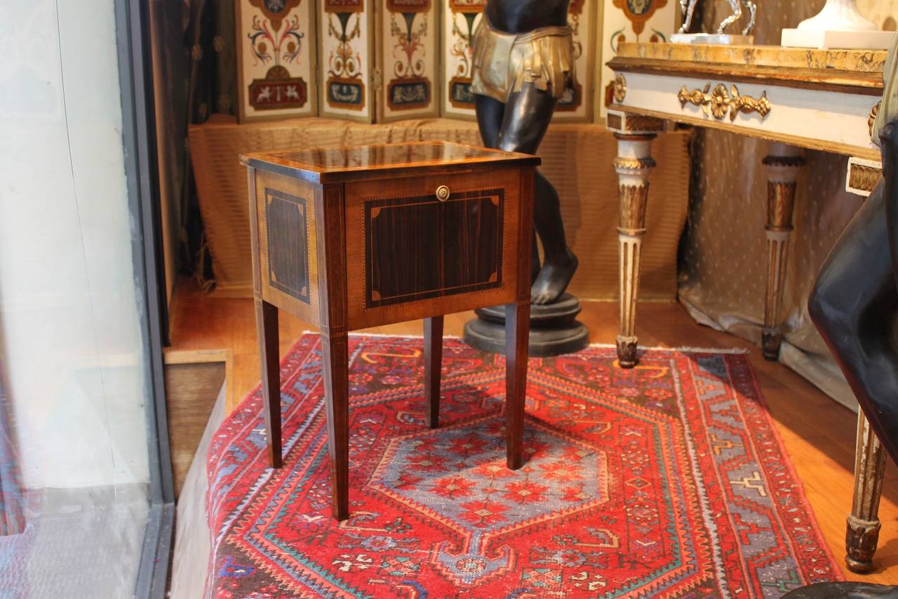 This fine late 18th century Italian side table or nightstand is made of flamed solid and veneered mahogany and walnut wood raised on four squared tapering legs.
Both the top and the sides are embellished with a very well executed marquetterie inlay