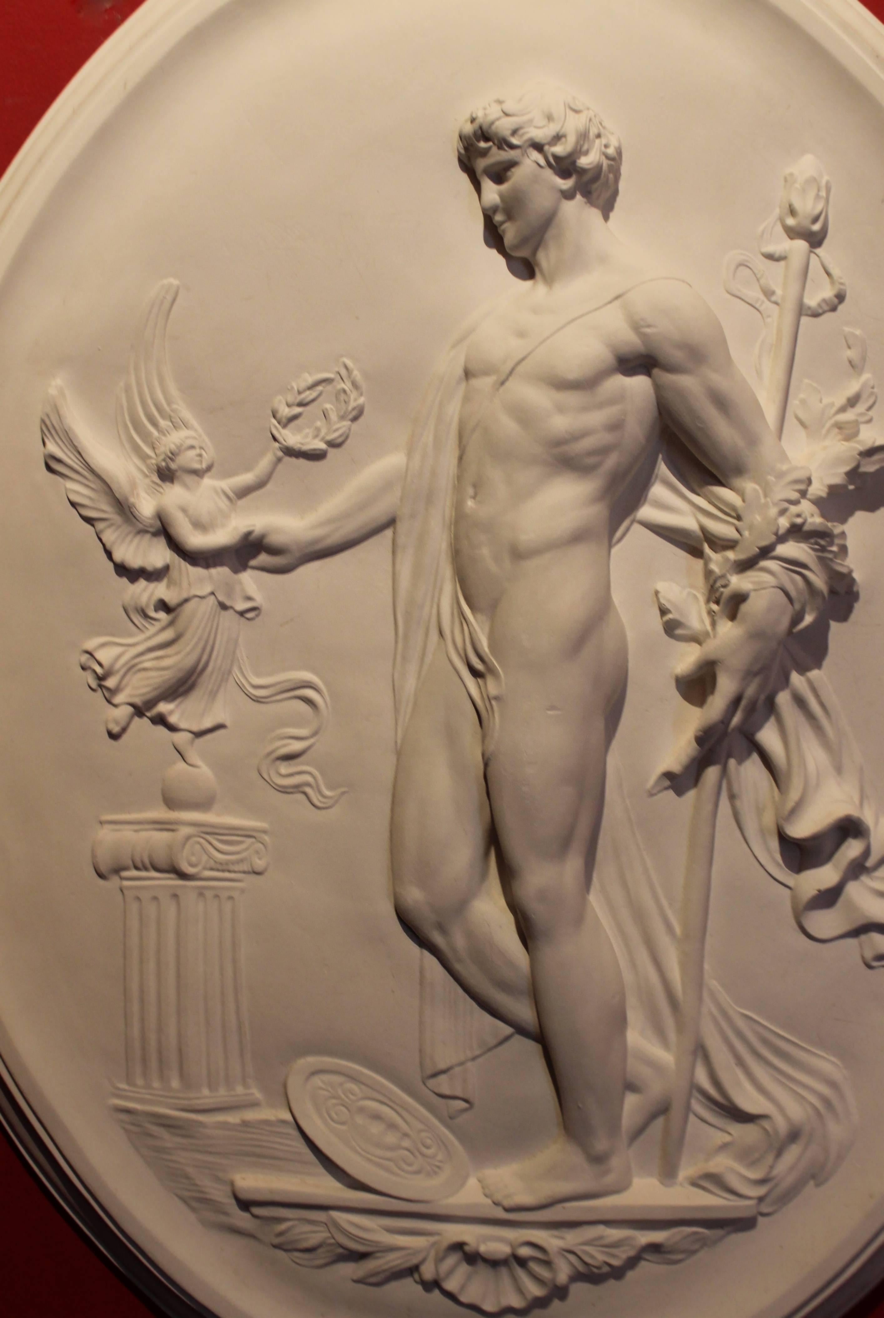 This late 19th century large white oval high relief plaster plaque represents an allegorical and mythological scene. The  male figure, Dionysus or Bacchus with the body covered by a drape, holds a wand (thyrsus) in his hand, his shield is leaning on