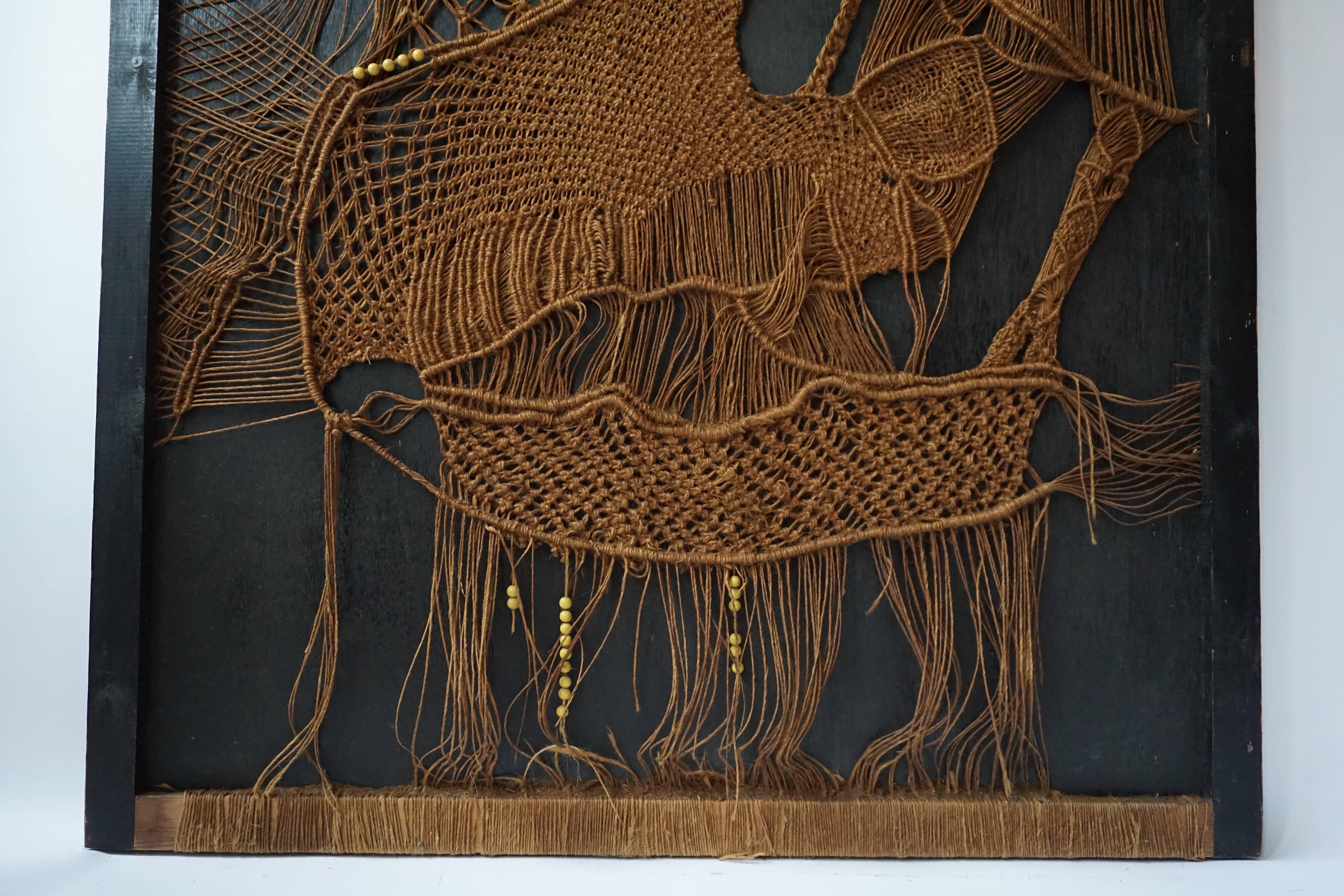 A 1960s–1970s so – called ‘ macramé ‘ panel of woven and beaded rope tapestry, representing an abstract pattern slightly reminiscing the figure of a bird, on a black painted wooden background.
Dimensions: 100 x 155 cm.

Some ropes are snapped