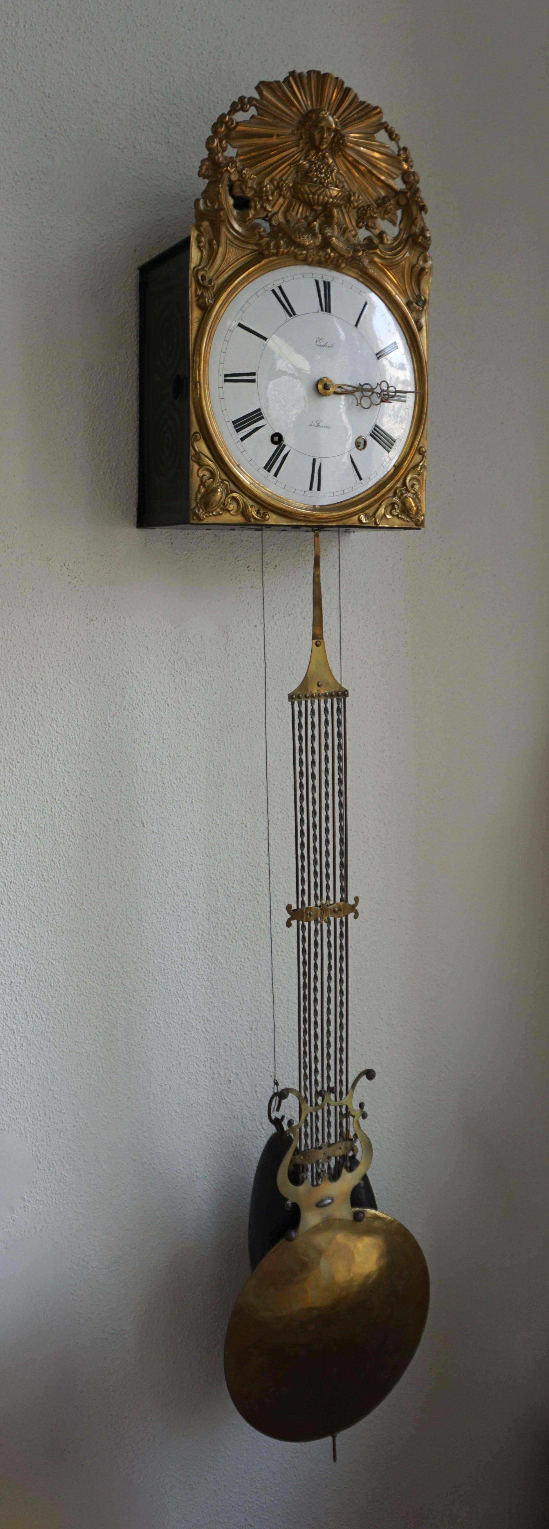 This two weight driven clock shows a white enameled dial with black Roman numerals surrounded by an embossed brass facade. The pendulum is also in brass and in lyre shape.

The clock strikes the hour-two times within two minutes and also the half