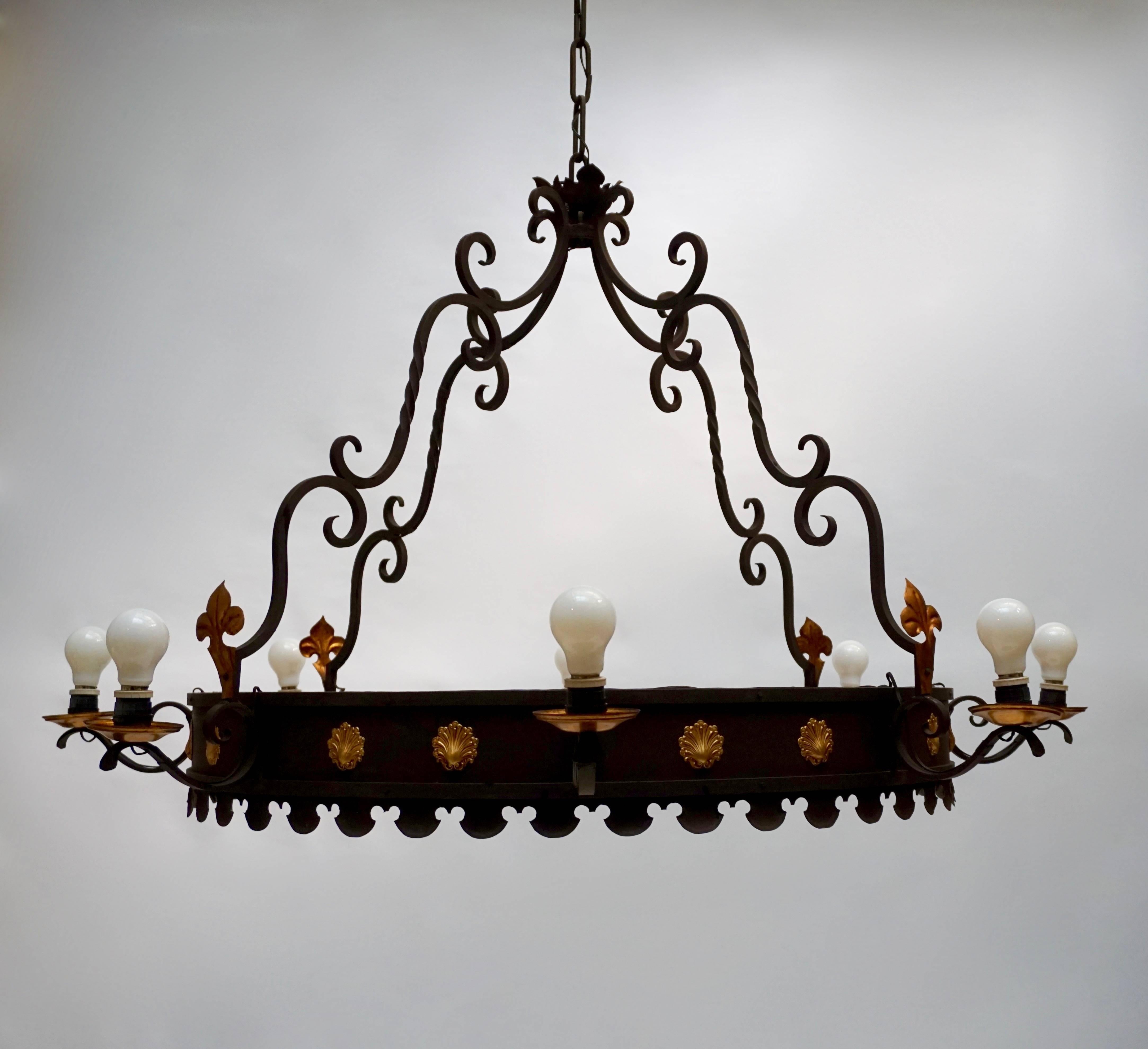 A very large and highly decorative wrought iron eight-light circular chandelier in the medieval revival style of the 1940s, decorated with gilt iron scallops, rosaces and lilies.

The chandelier has eight sockets for incandescent lamps with screw
