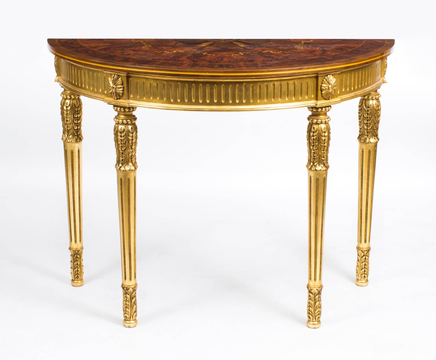 This is an exquisite pair of Sheraton style giltwood half moon console tables with elaborate marquetry inlaid decoration dating from the late 20th century. 

Sheraton is a style of English furniture that originated around 1800. This style is