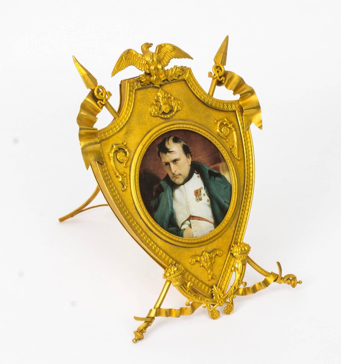 This is a lovely antique French Empire Revival gilt bronze picture frame, circa 1880 in date. 

The shield shaped frame features a spread eagle crest with military flags and laurel wreaths on either side. The ribbon wrapped Stand is joined by a