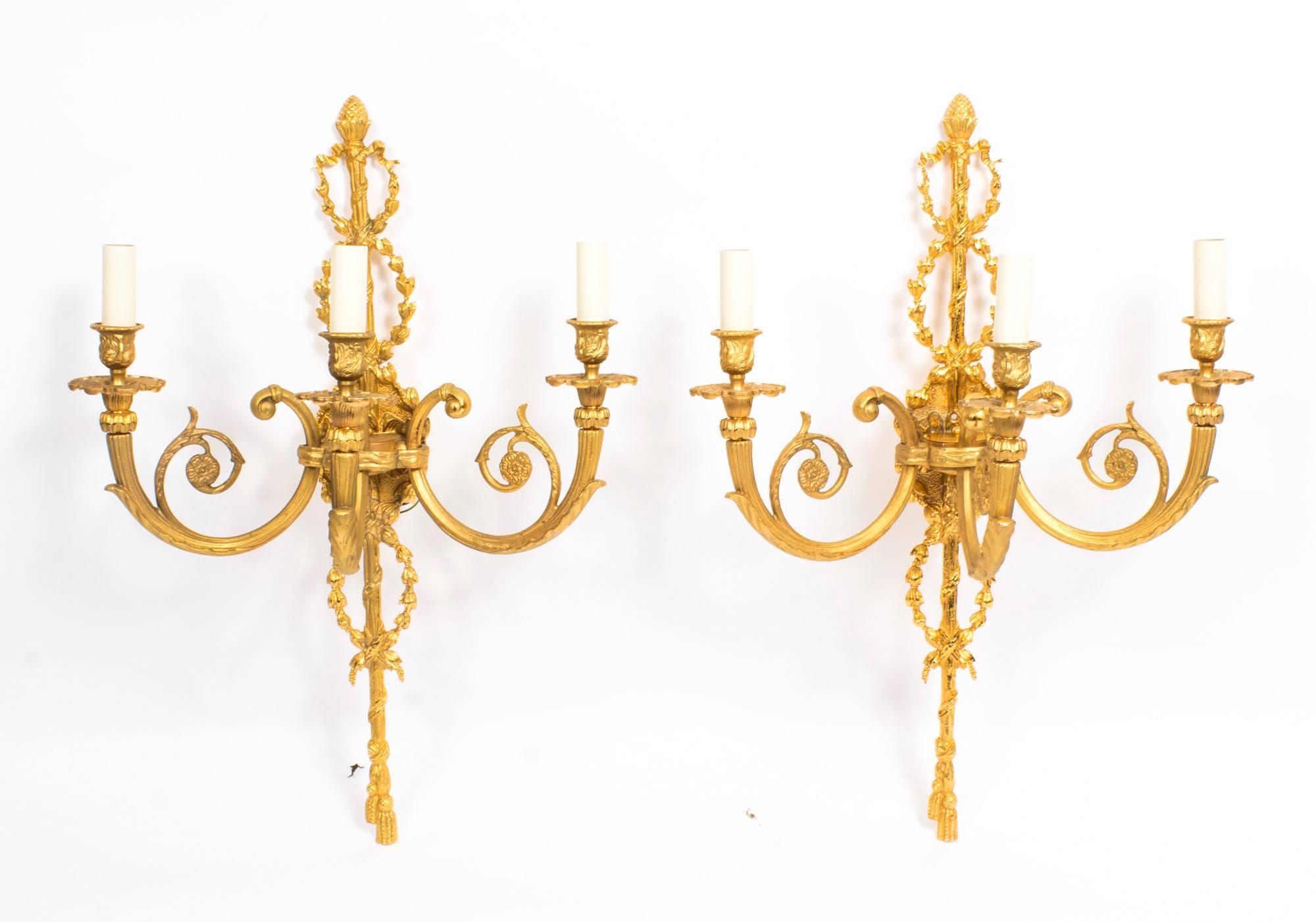 This is a stunning pair of vintage French neoclassical ormolu gilt bronze three branch wall sconces, circa 1920 in date.

They feature ribbon tied and torch form back plates with laurel swags, scrolling branches with floral ends and urn form