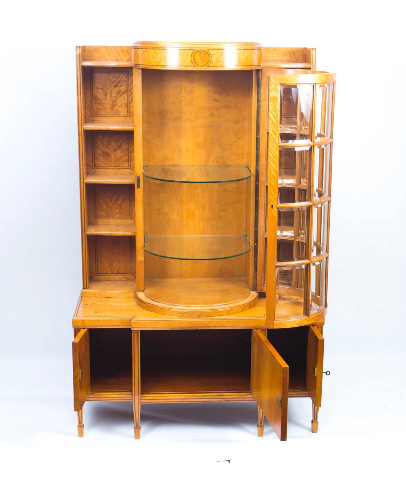 This is a stunning antique late 19th century satinwood display cabinet, outlined throughout with boxwood and ebony stringing. It has a decorative central bowed door with twenty bevelled glass panels, opening to reveal two adjustable glass shelves.