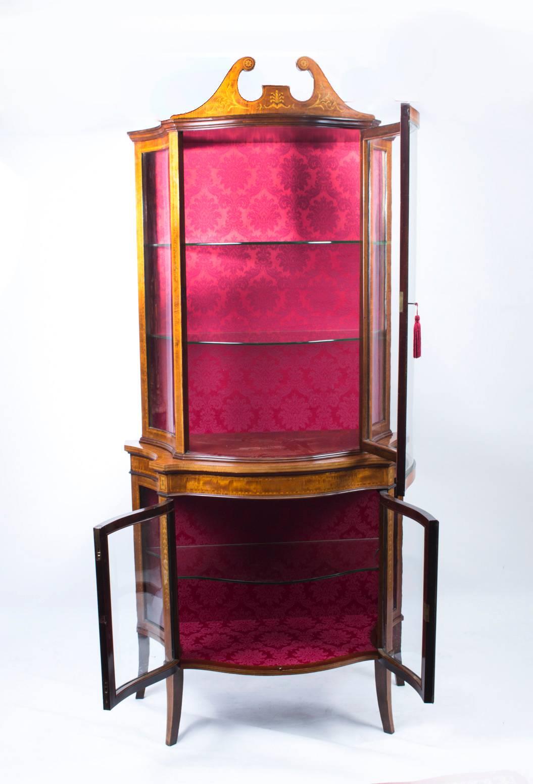 This is a stunning English Edwardian serpentine mahogany display cabinet, circa 1890 in date with exquisite hand cut inlaid decoration.

It has a glazed door above a pair of glazed doors, opening to a red damask lined interior with glass
