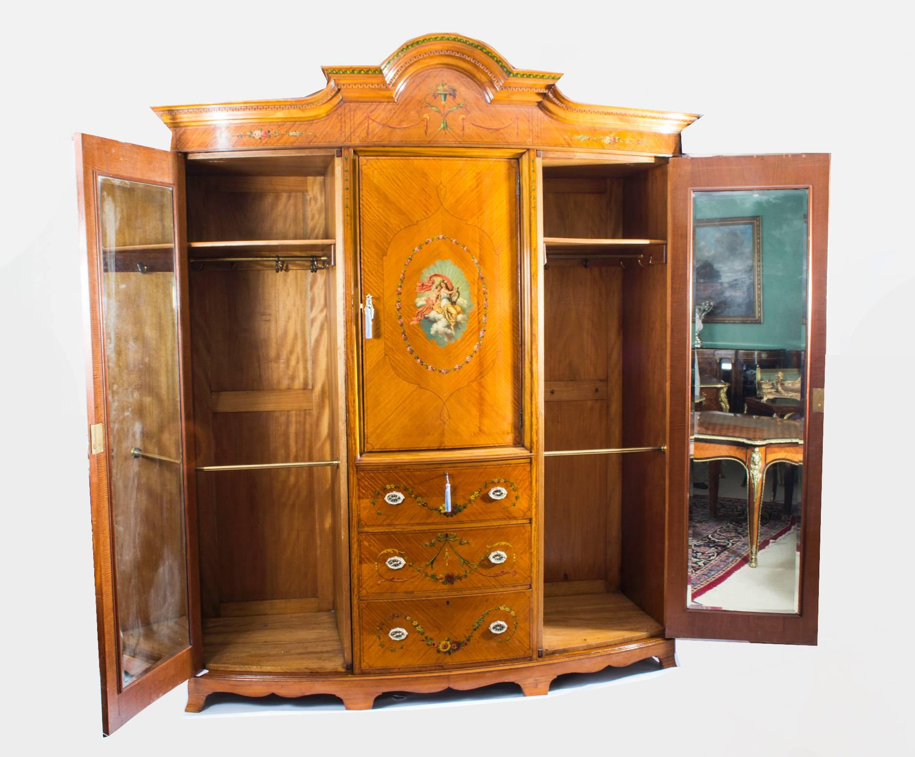 This is a large and impressive English antique Victorian satinwood and hand-painted bow-front wardrobe, circa 1880 in date.

It is of neoclassical design and is beautifully painted, in the manner of Angelica Kauffman with a basket of fruit, musical