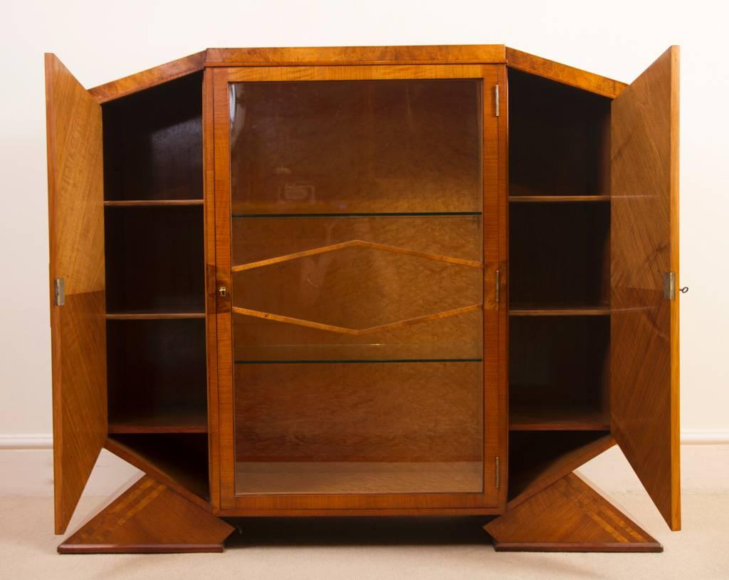 This is a beautiful antique Art Deco satinwood maple and walnut display cabinet, circa 1920.

The cabinet is almost octagonal in shape and stands on two elegant triangular feet.

The side doors open to reveal interiors finished in walnut each