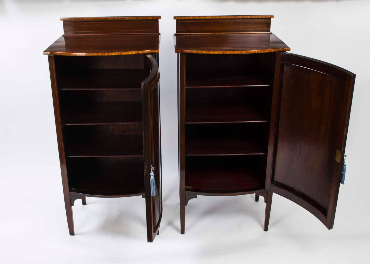 This is a lovely pair of antique Edwardian bow fronted mahogany music cabinets, circa 1900 in date.

The pair has raised backs, feature decorative satinwood inlay of musical instruments to the doors and stand on square tapering legs. 

Each door