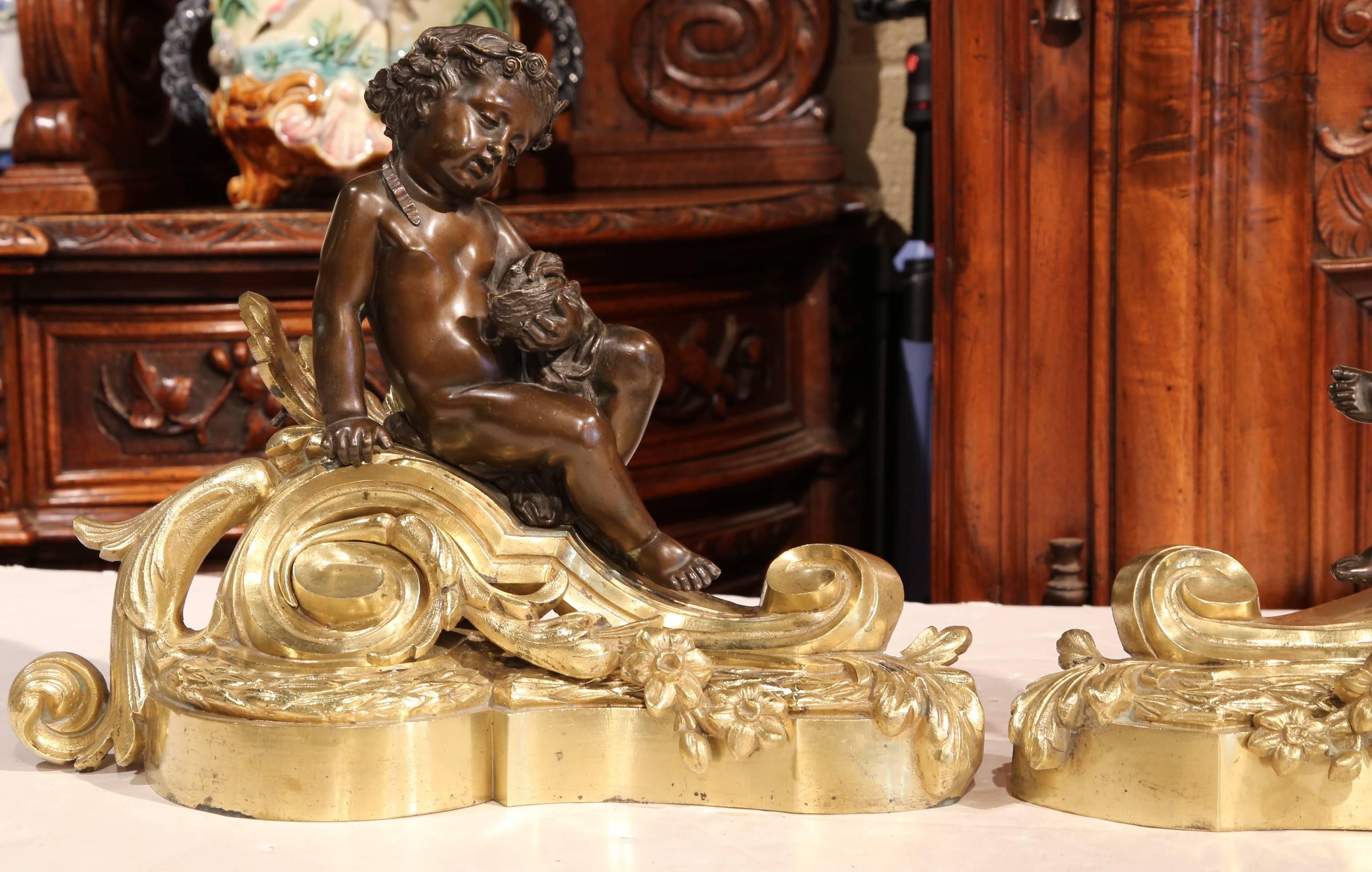 Decorate your fireplace with this elegant pair of antique patinated bronze andirons chenets. Crafted in France, circa 1840, the fireplace essentials feature a pair of cherubs seated and draped over a scrolled foliate stepped base; each cherub has