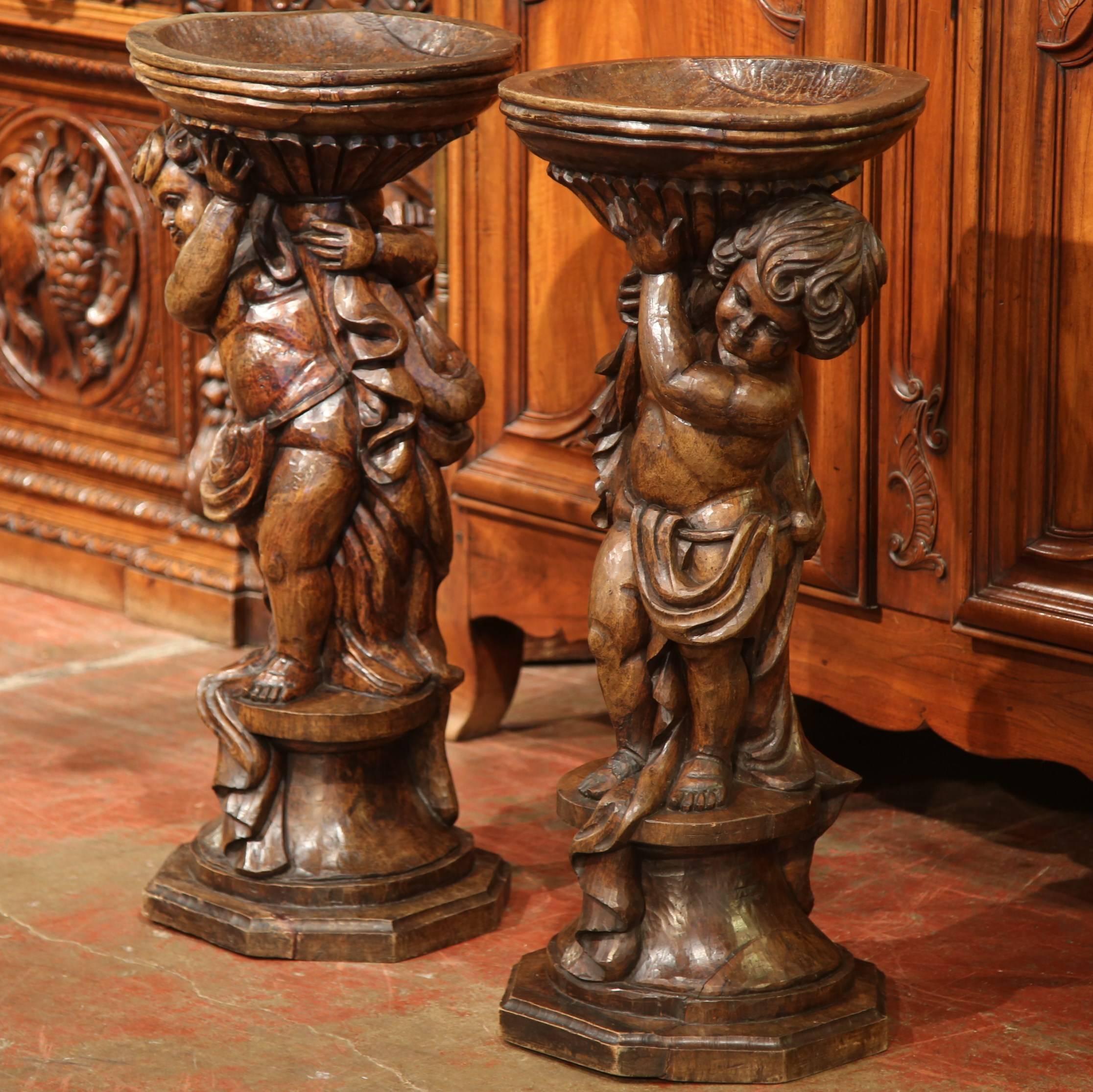 This elegant pair of antique planters was created in France, circa 1790. The base of the stands are shaped as a pair of cherub figures, who hold up shallow, round jardinière bowls for plants. Both ornate, detailed wooden pieces are hand-carved from