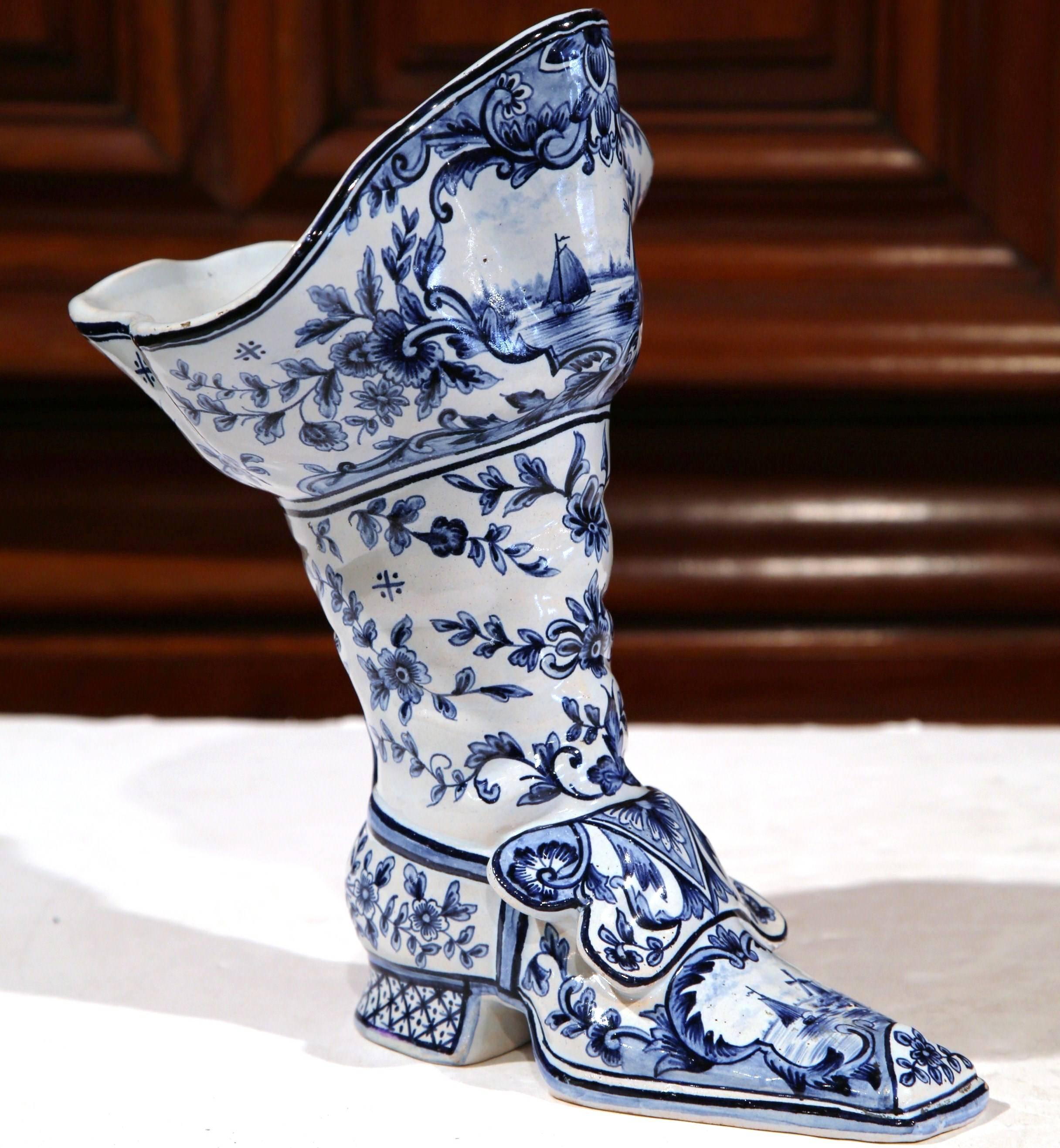 Decorate a tabletop or surface with this interesting faience vase. Crafted, circa 1900, the colorful vase is sculpted to be a man's boot, and embellished with hand-painted blue and white delft hand-painted motifs including a windmill, sailboat and