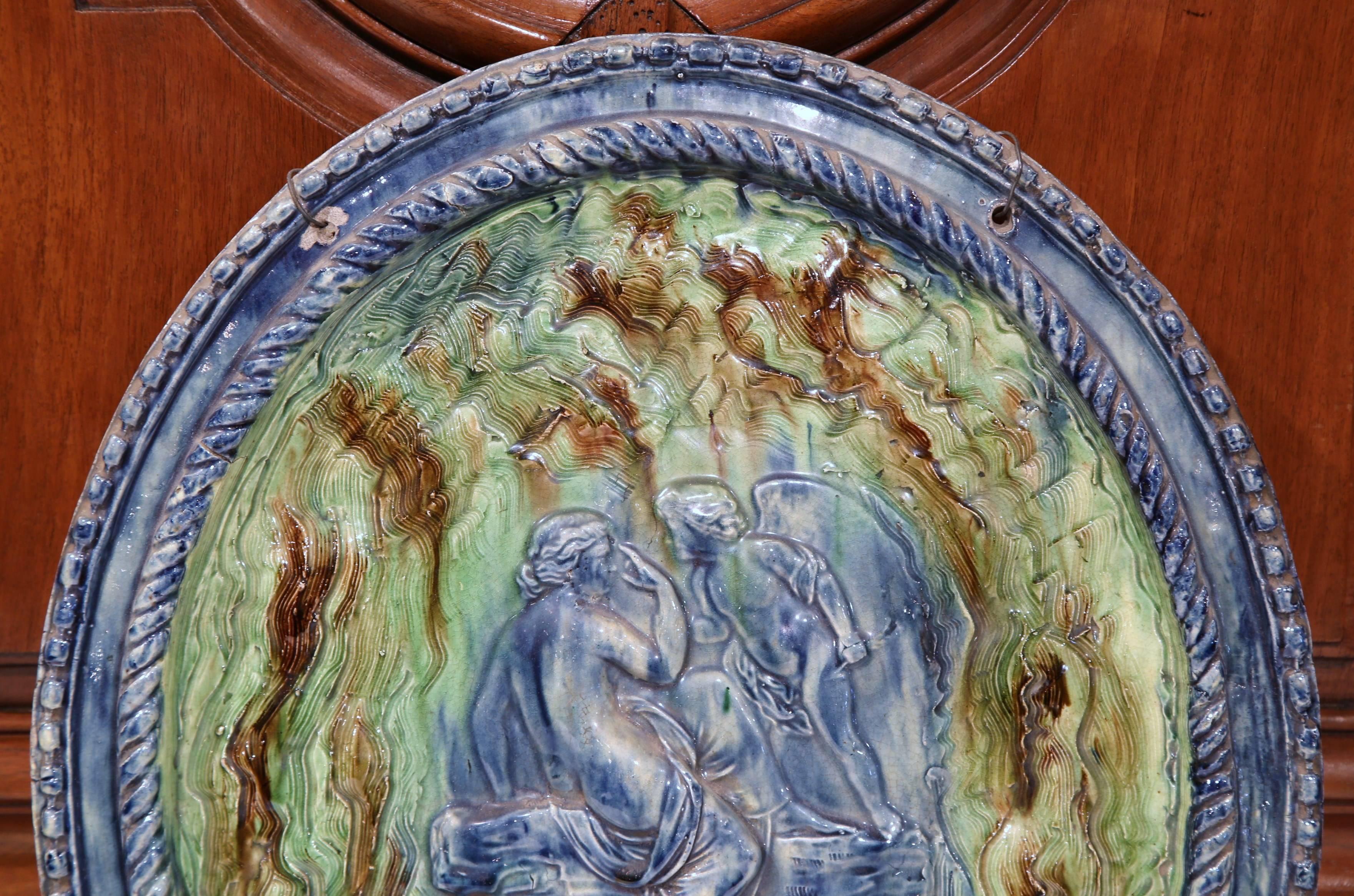 Cette plaque de suspension colorée, en terre cuite ancienne, a été sculptée à Beaune, en France, vers 1860. L'assiette ovale est peinte à la main dans une palette de vert et de violet et présente une belle texture en trompe-l'œil. Cette décoration