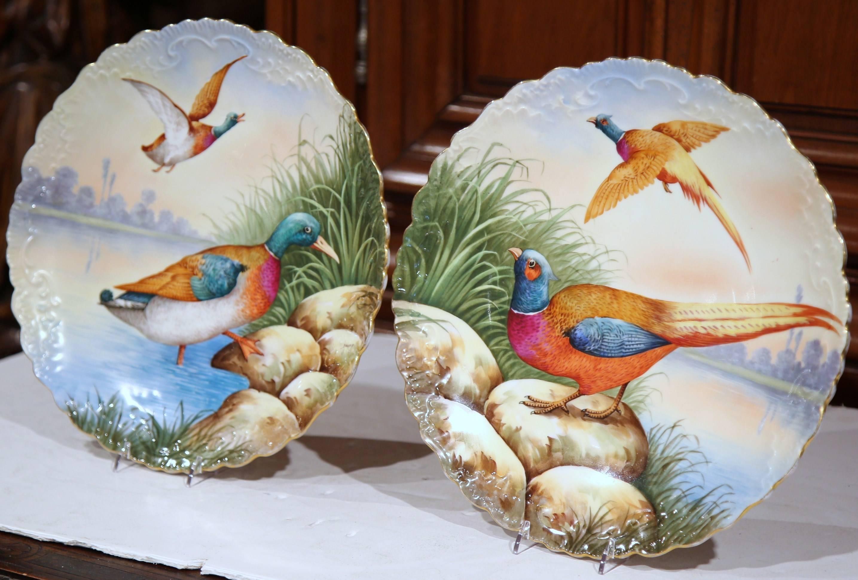 This large, colorful pair of antique porcelain wall hanging platters were sculpted and painted in Limoges, France, circa 1880. Both circular, ceramic plates have scalloped edges with gold trim and are hand-painted; two ducks on one and a pair of