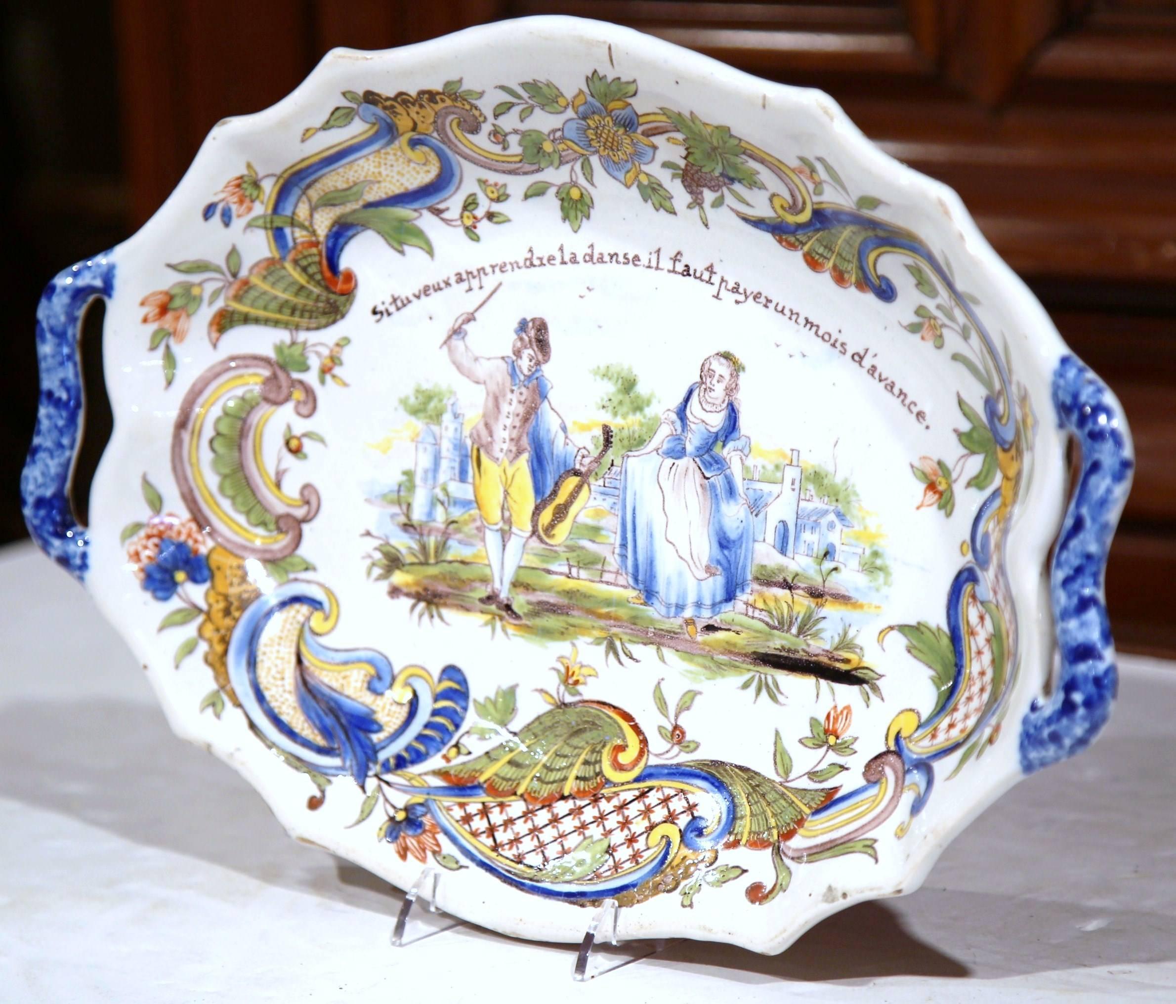 This beautiful antique ceramic dish was crafted in Normandy, France, circa 1880. The stylized ceramic plate has a scalloped edge and depicts a hand painted courting scene with a violinist teaching a young woman how to dance. The French motto reads: