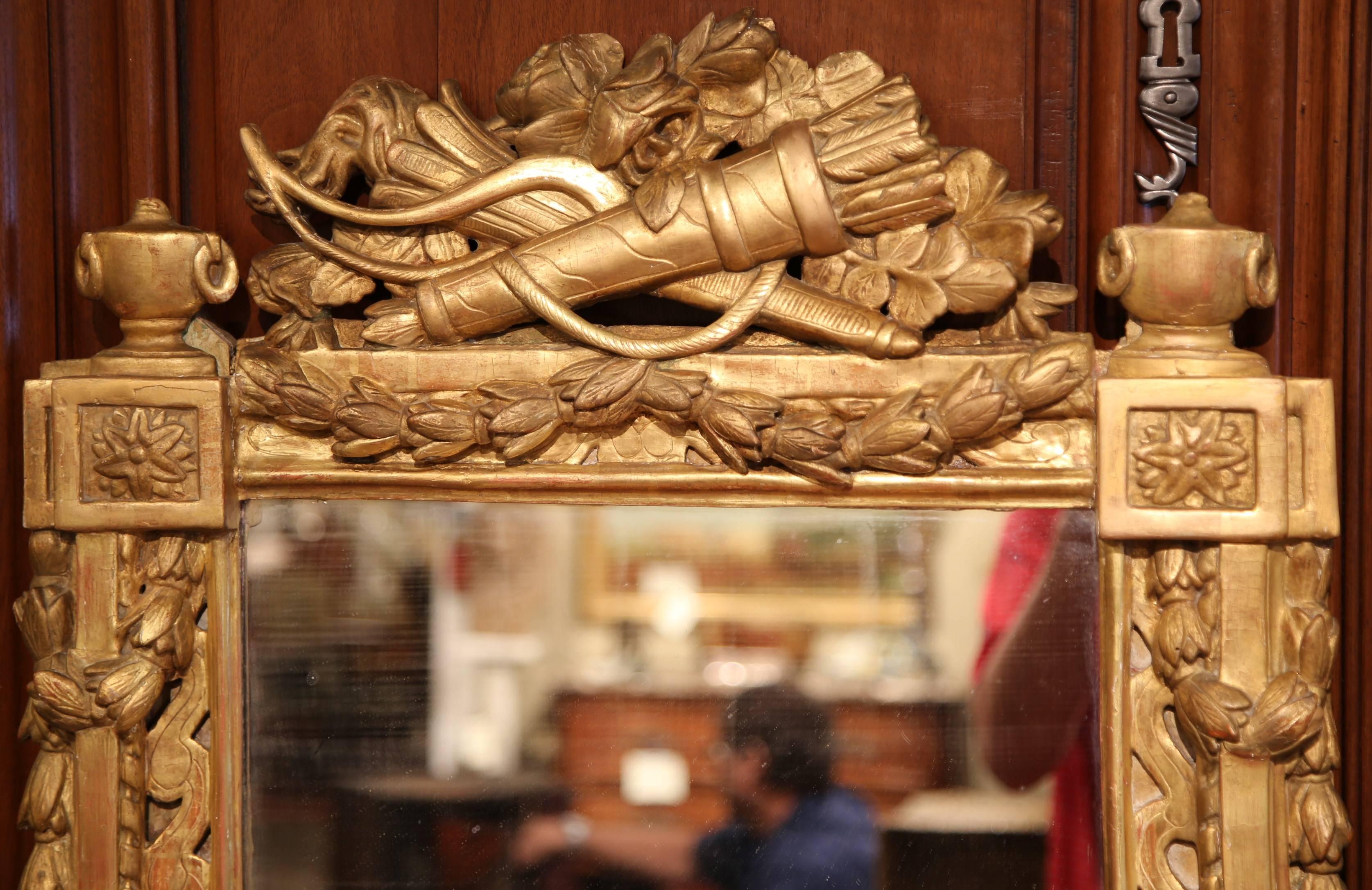 This elegant antique gilt mirror was crafted in Southern France, circa 1780. The wall mirror is heavily carved; the pediment is formed by a quiver of arrows, crossed by a pair of torches, and further embellished with a classic ribbon of laurel