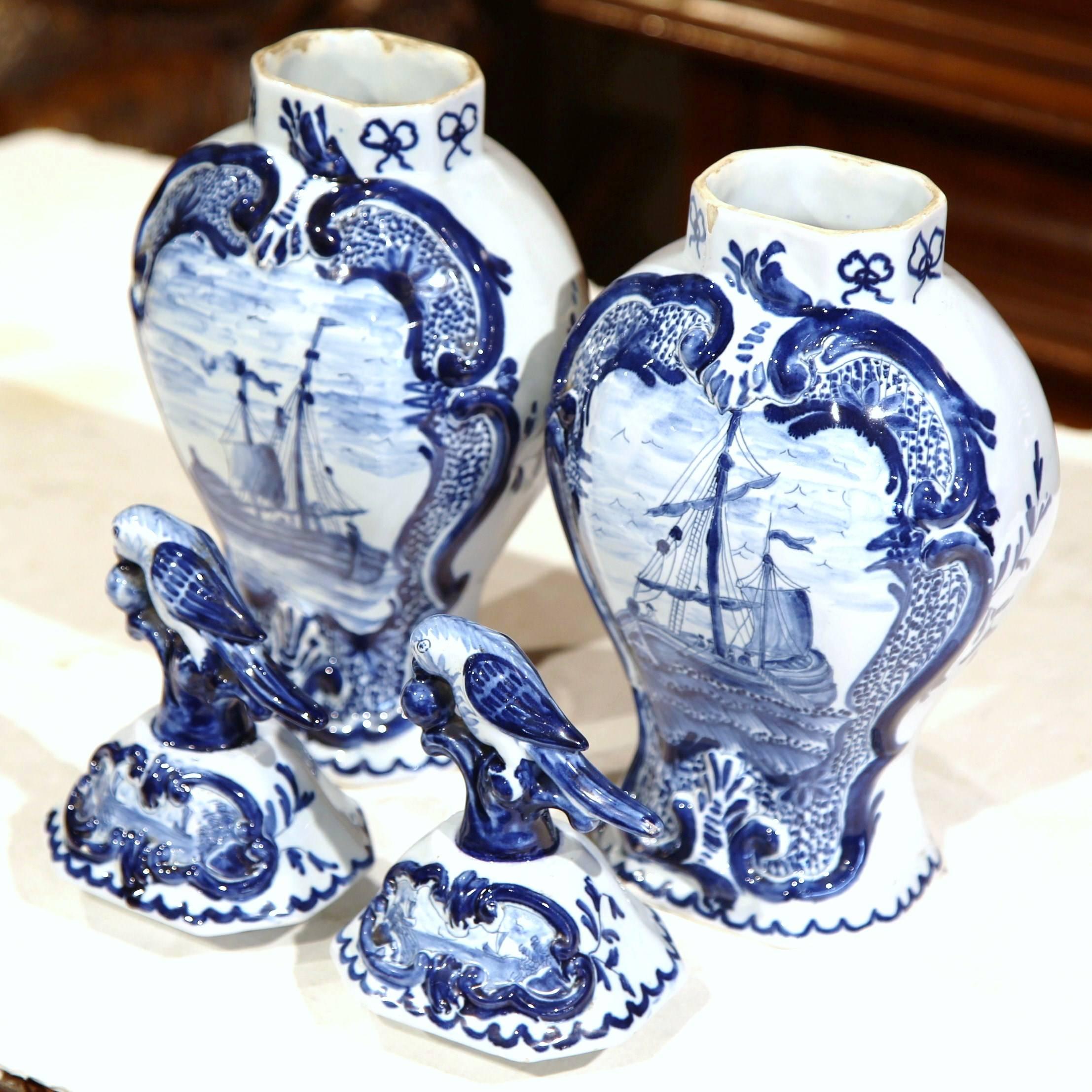 This elegant pair of faience vases were sculpted in France, circa 1920. Created in the manner of Delft, both of the classic blue and white vases feature maritime sailboat and lids with a parrot picking a cherry. The porcelain pieces are signed on