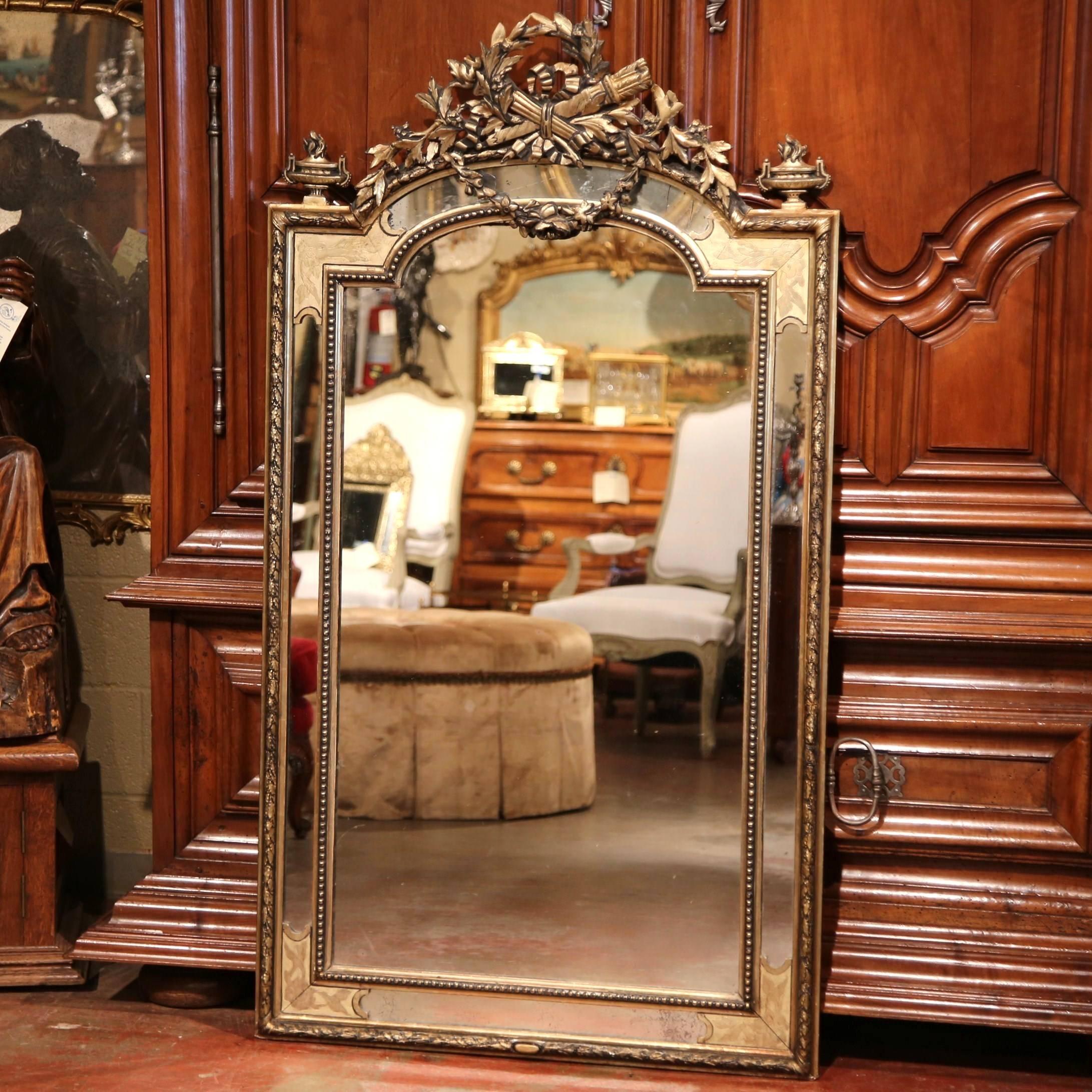 This elegant, antique wall mirror was crafted in Paris, France, circa 1870. The intricate, carved pediment features a Classic, iconographic motif, with two crossed torches and the traditional Louis XVI ribbon bow inside a laurel wreath, embellished