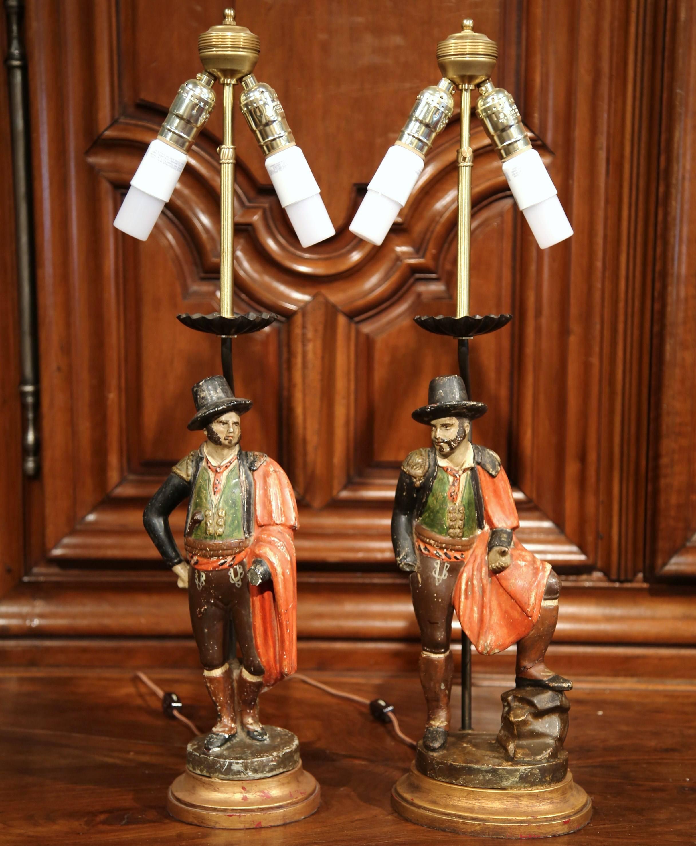 This beautiful pair of lamps was crafted in Spain, circa 1870. Standing on a gilt wooden base, each lamp depicts a carved matador figure with a red cape on his shoulder. Both light fixtures have their original painted finish in a red and brown color