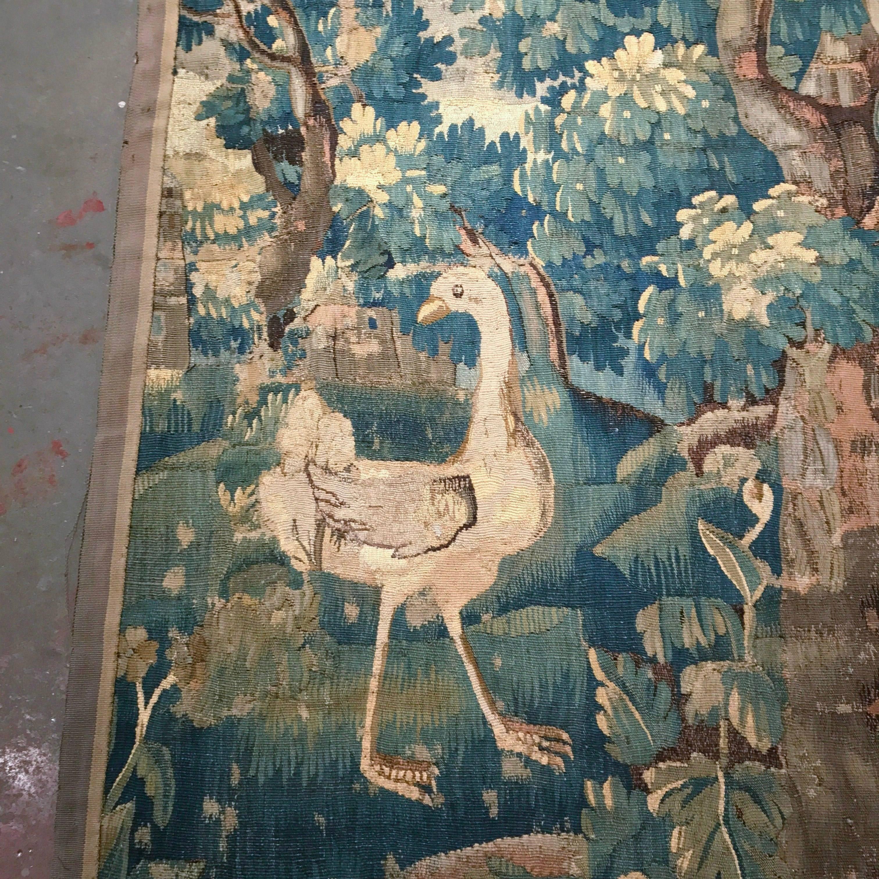 This colorful, antique tapestry was woven in Aubusson, France, circa 1750. The rectangular, wall hanging verdure tapestry, features a tall ostrich in the foreground in front of a large tree filled with heavy foliage, and is further animated with a